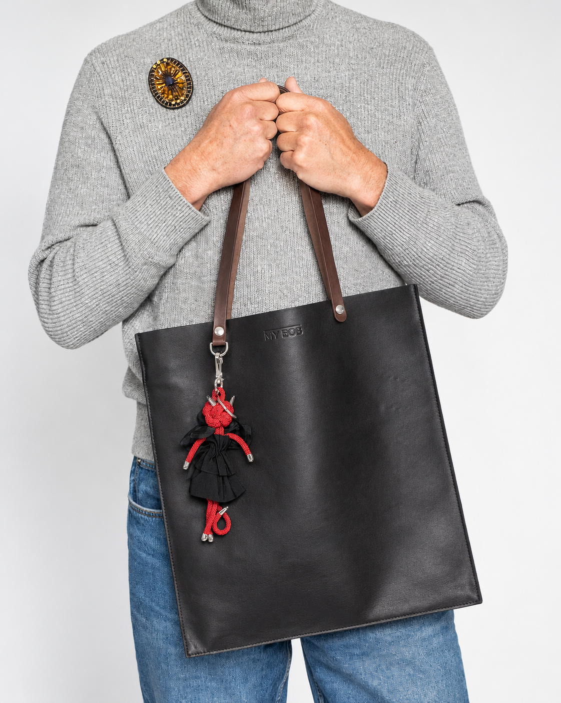 A person wearing a gray turtleneck sweater and blue jeans holds a large black leather tote bag with leather straps. The bag has a small logo plate and a red-and-black dangling charm attached near the top.