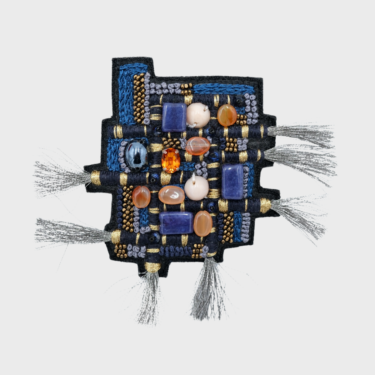 A square black embroidered brooch featuring blue, purple, white, and orange beads, small gold-tone rings, and gray tassel fringe ends, shown on a light gray background.