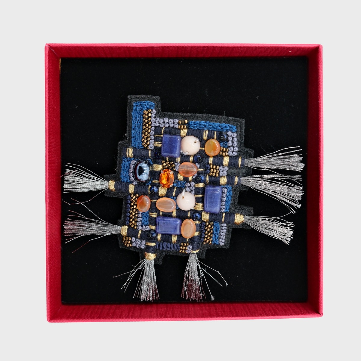 A square decorative brooch with blue, black, white, and orange woven details and silver tassel ends, presented inside a red square box with a black lining on a light gray background.