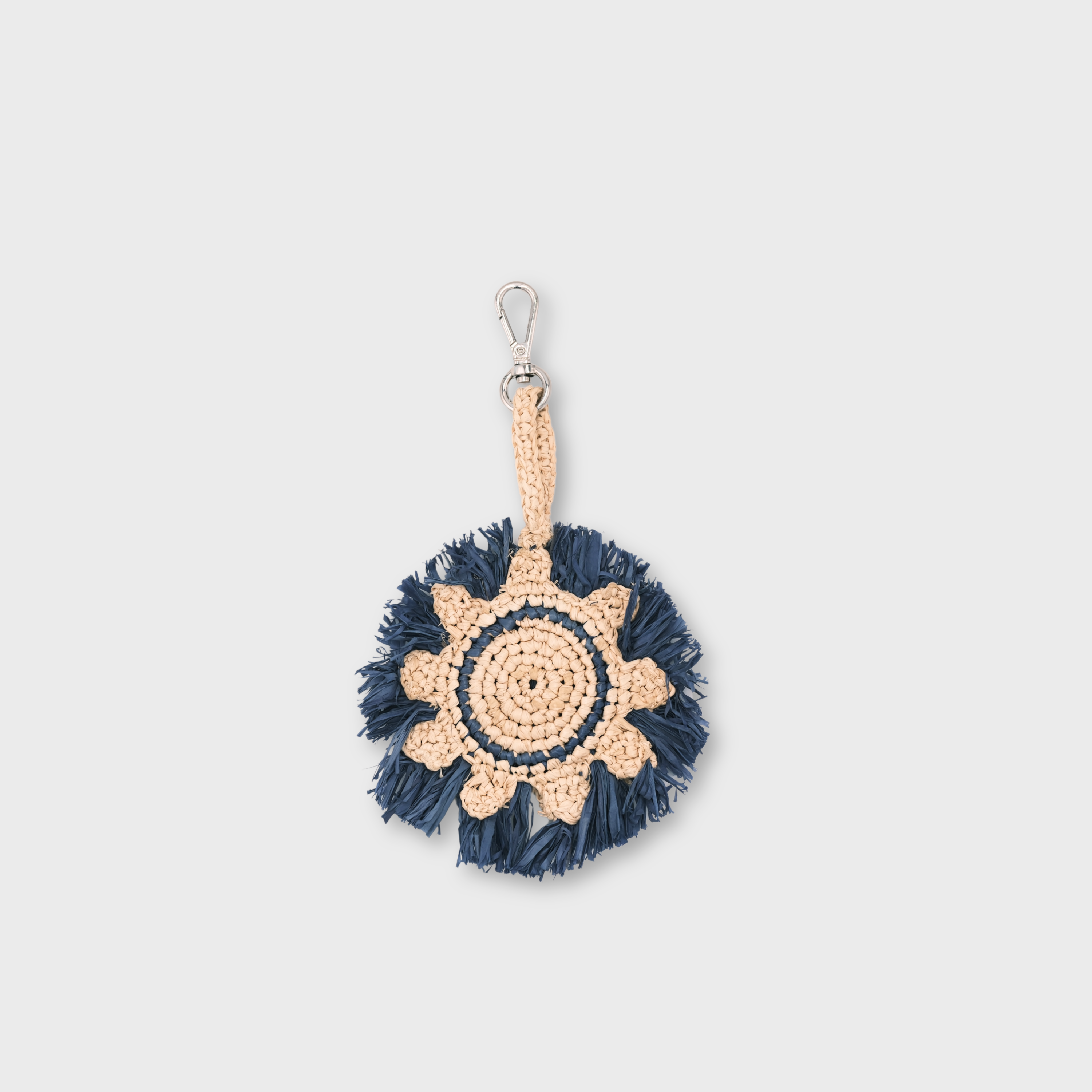 A handmade raffia keychain featuring a circular pattern in natural beige and dark navy blue, representing the sun, finished with a silver metal lobster clasp at the top.