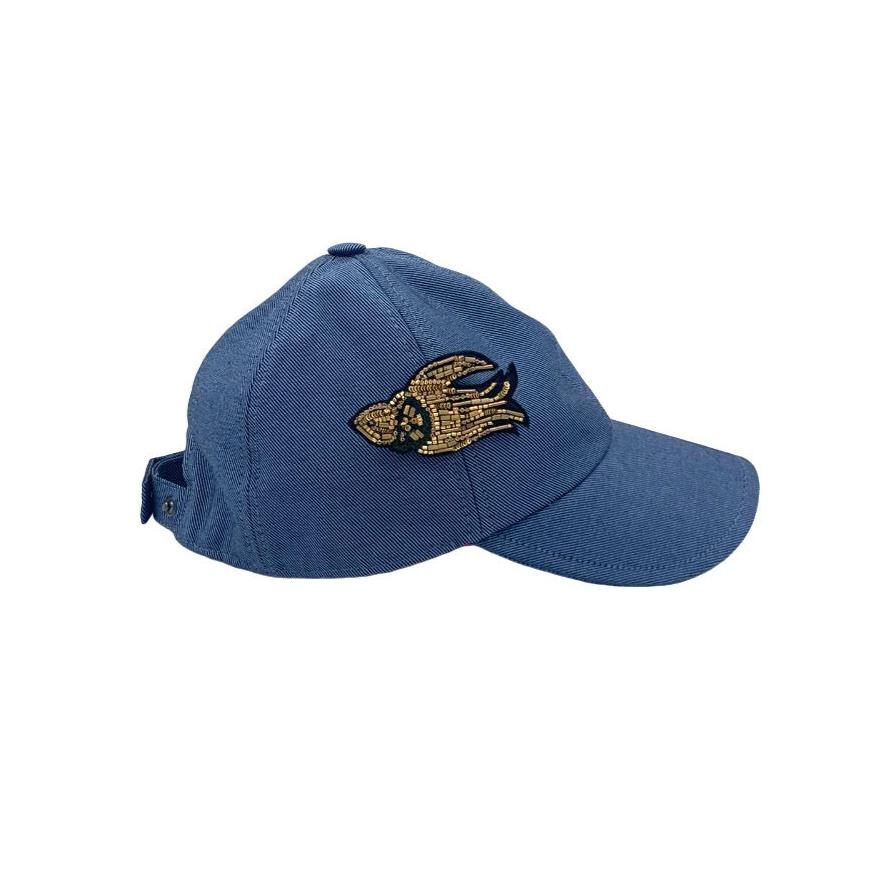 A blue jean cotton cap from the side, with a golden embroidered brooch representing a bird.
