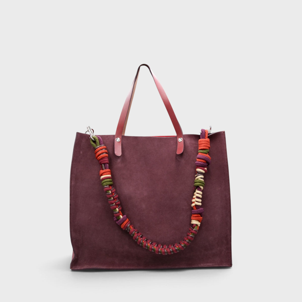 A large burgundy suede tote bag with reddish-brown leather handles and a thick braided macrame shoulder strap featuring orange, red, green, and cream woven accents, shown against a light gray background.
