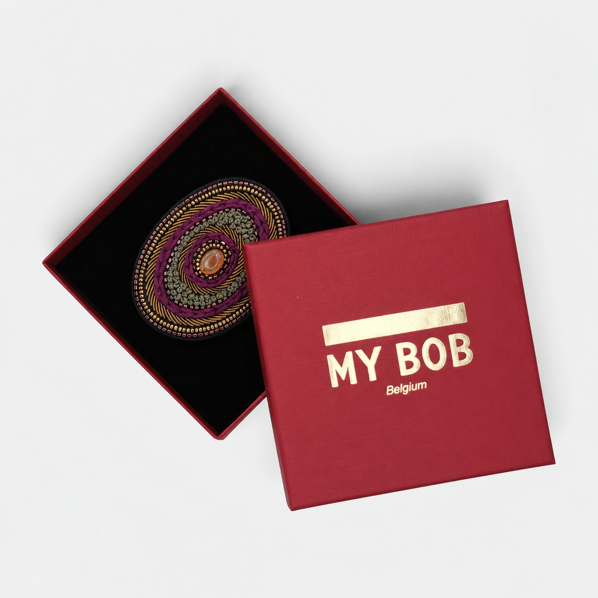 A circular, hand-embroidered brooch with concentric patterns in purple, olive green, gold, and black, featuring a small orange and white bead-like center shown in a red box with black velvet.