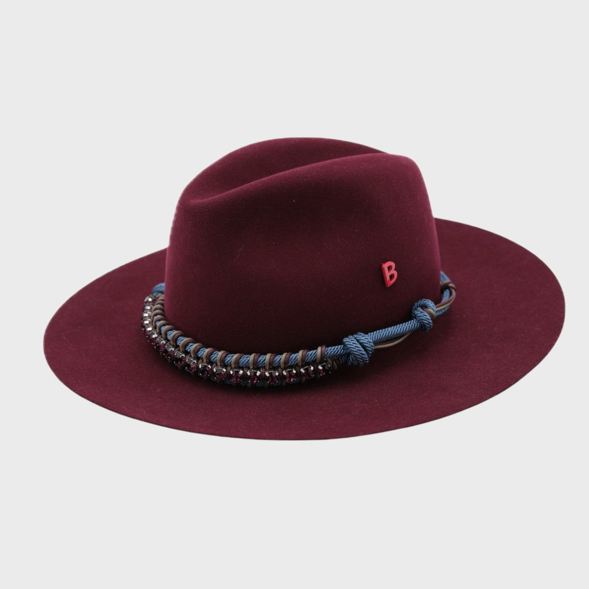 Burgundy fedora hat with decorative band with purple rhinestos and blue macramé and red 'B' pinned on it on a light gray background