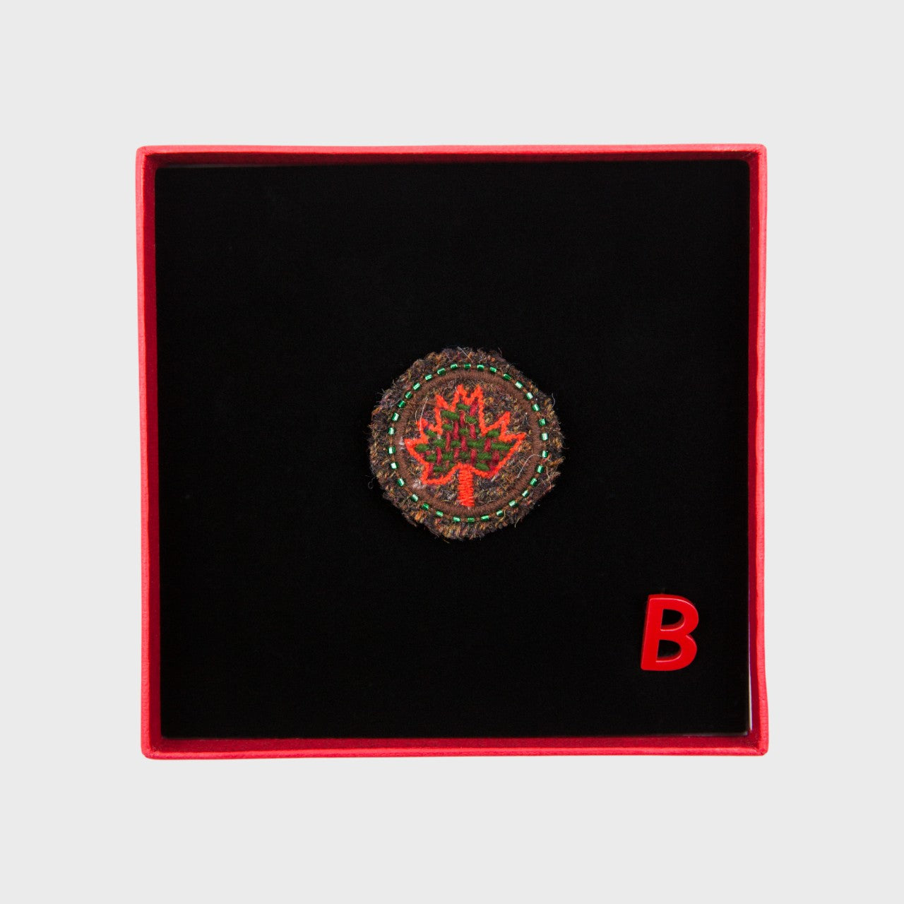 An embroidered brooch representing the red canada leaf in a box.