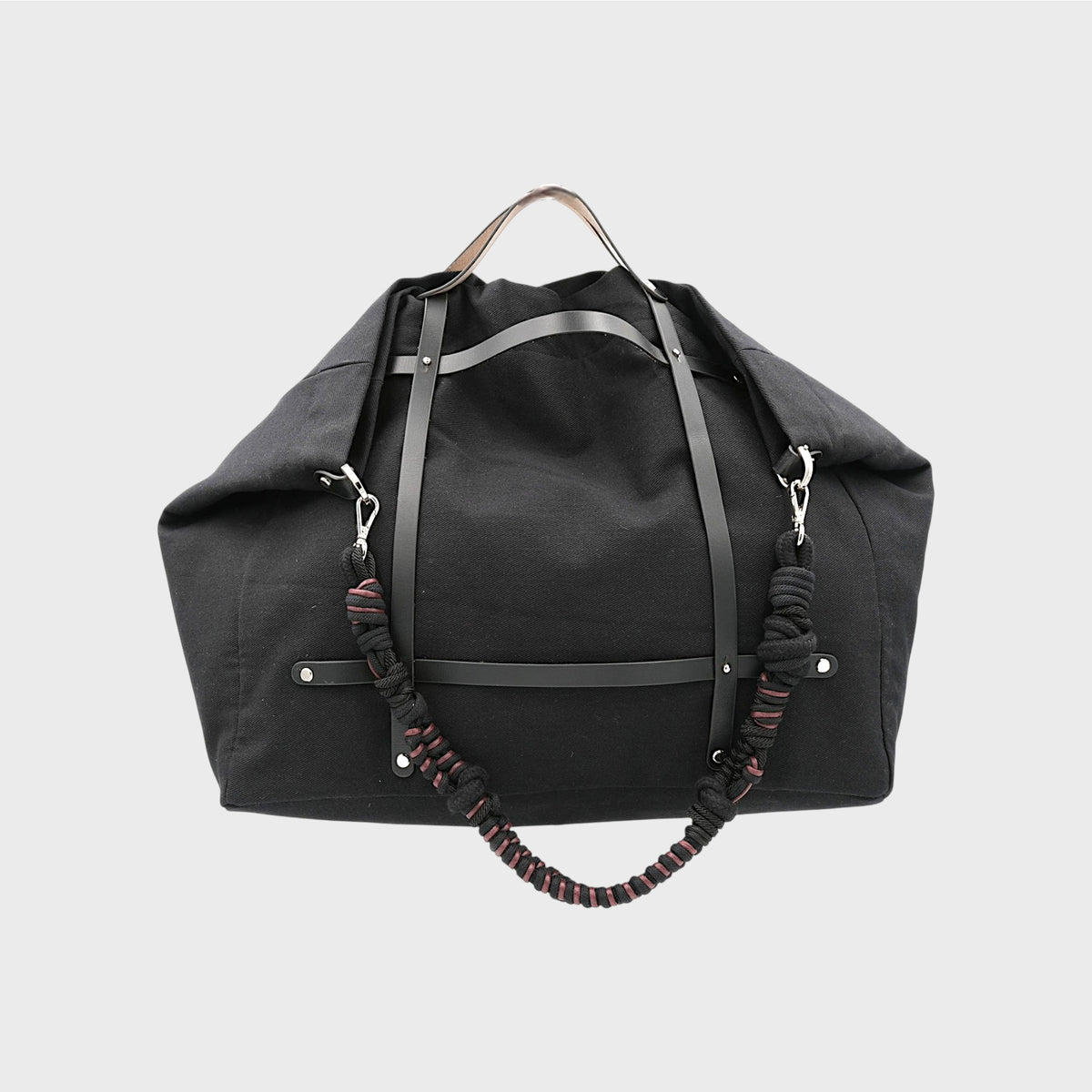 Black duffel bag with leather straps and a black and brown macrame shoulder strap on a light gray background.