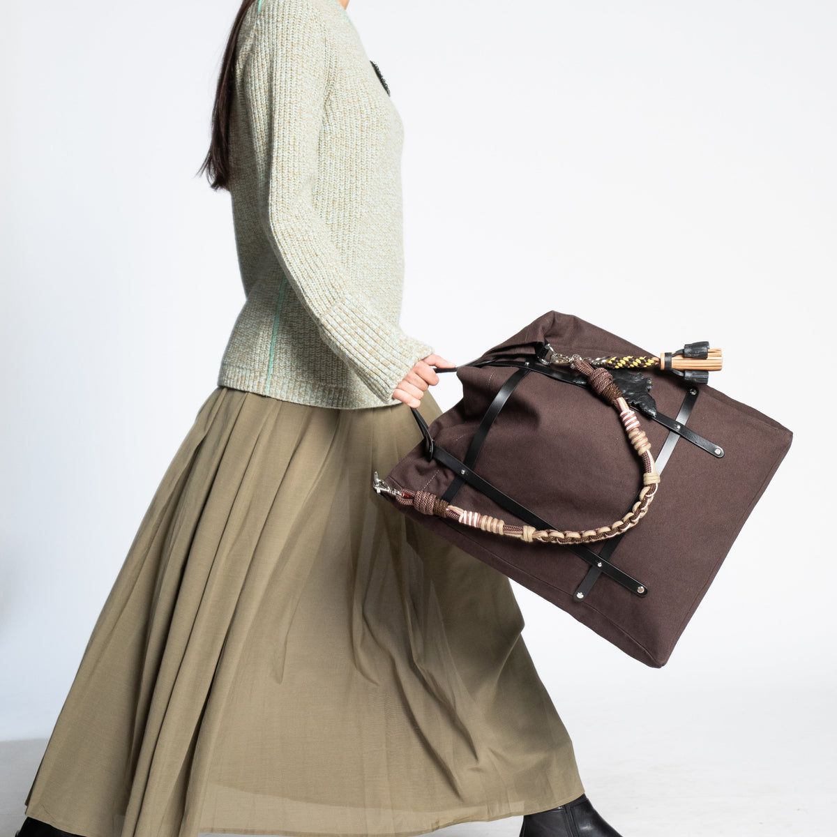 A woman walks while carrying a chocolate canvas tote bag with a patterned strap, against a light gray studio background.