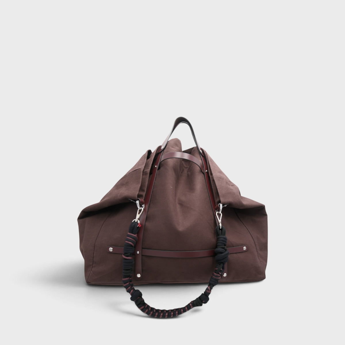 A brown canvas duffel bag with dark brown leather trim and a black-and-brown knotted rope-style shoulder strap, photographed against a light gray background.