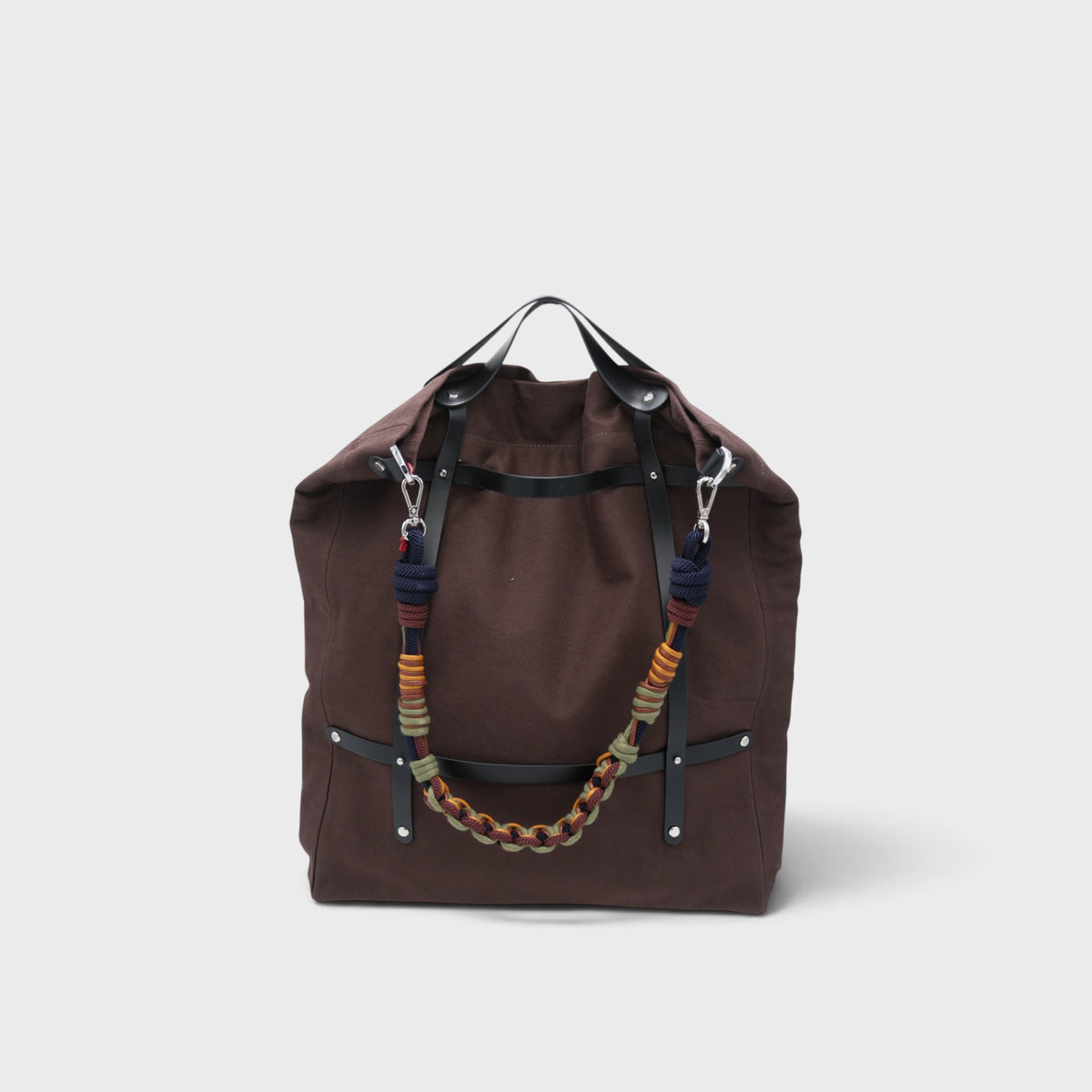 A canvas tote bag in a chocolate color, featuring brown leather straps and a decorative shoulder strap with blue, green, yellow details.