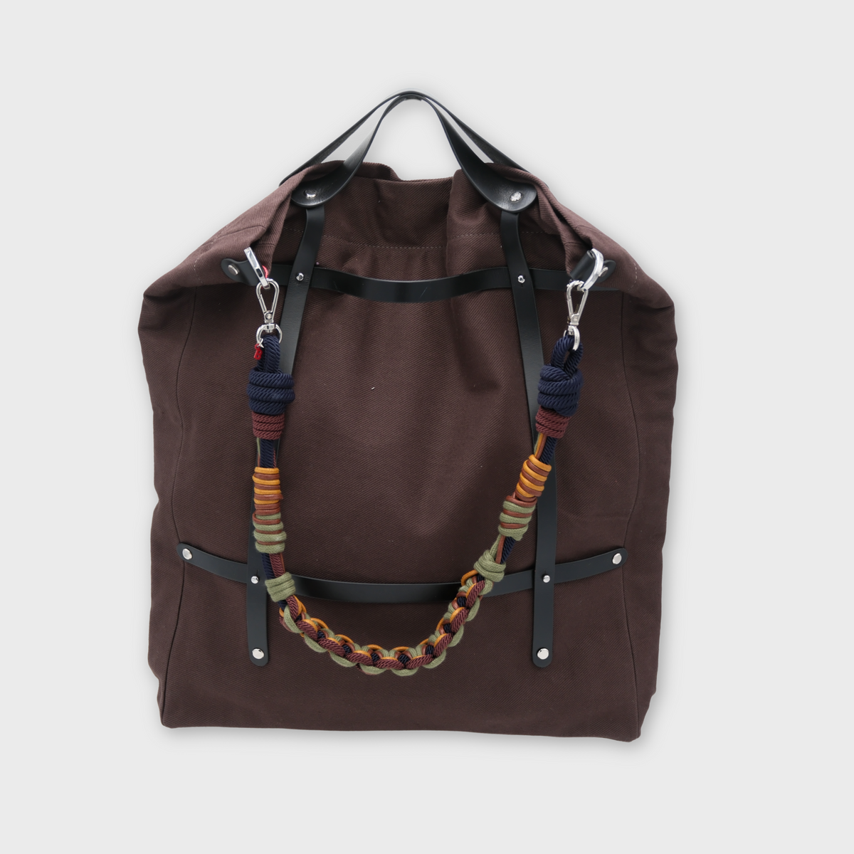 A canvas tote bag in a chocolate color, featuring brown leather straps and a decorative shoulder strap with blue, green, yellow details.