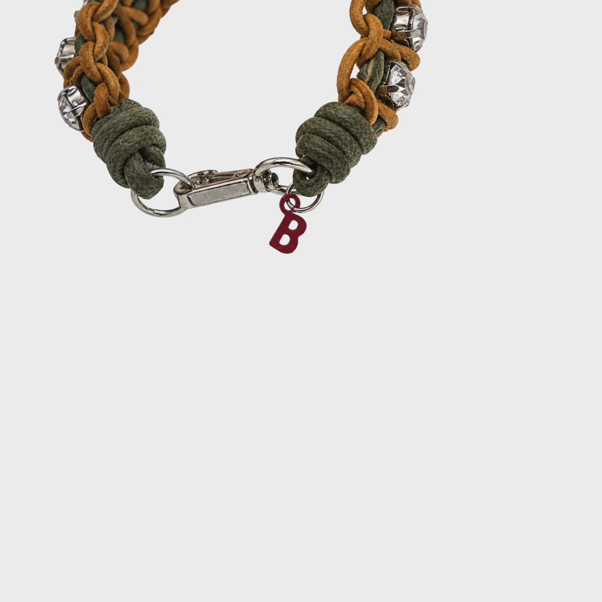 A close-up of a braided bracelet featuring olive green cord, tan cord, silver-toned metal links and rhinestones, and a small red letter 'B' charm, shown against a light gray background.