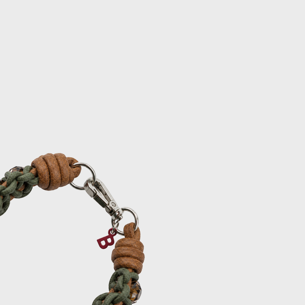Close-up of a braided macrame accessory with a green and brown woven cord, and a silver clasp with a small red letter B charm, photographed on a light gray background.