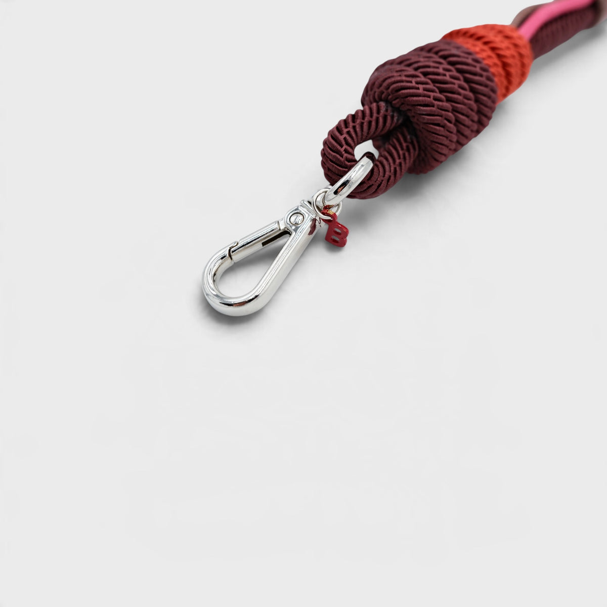 A close-up of a braided macramé bag strap with a silver metal clasp, featuring dark burgundy, rust orange, and pink braided cords on a light gray background.
