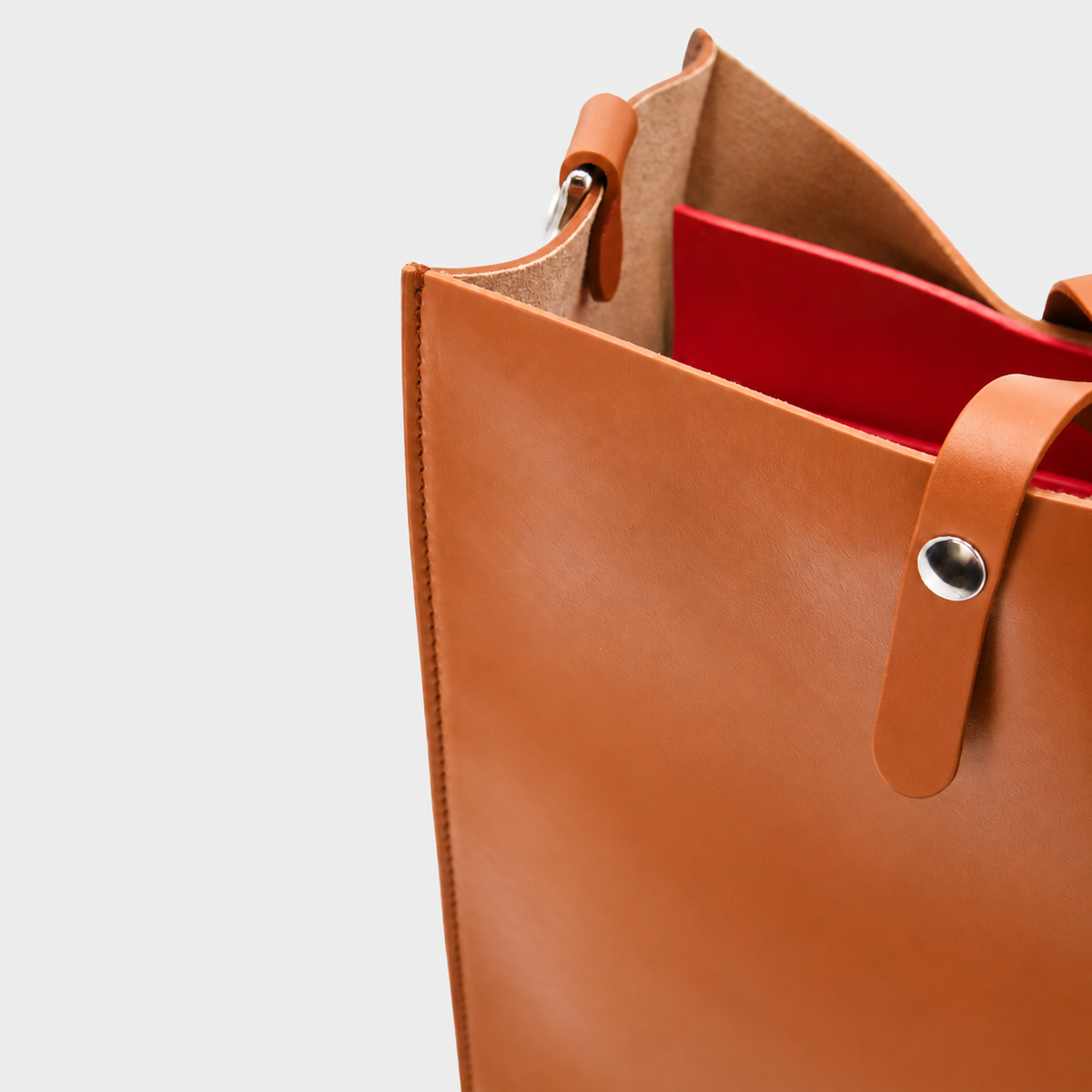 Close-up of a tan leather tote bag with a bright red interior lining, showing the strap attachment and metal rivet on a light gray background.