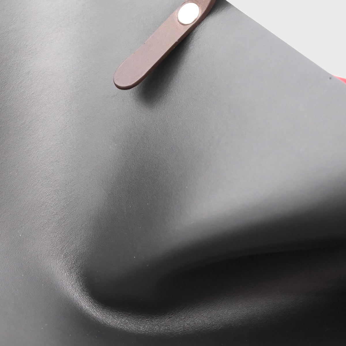 Close-up of a black leather bag with a brown leather strap and a silver metal rivet, shown against a light gray background.