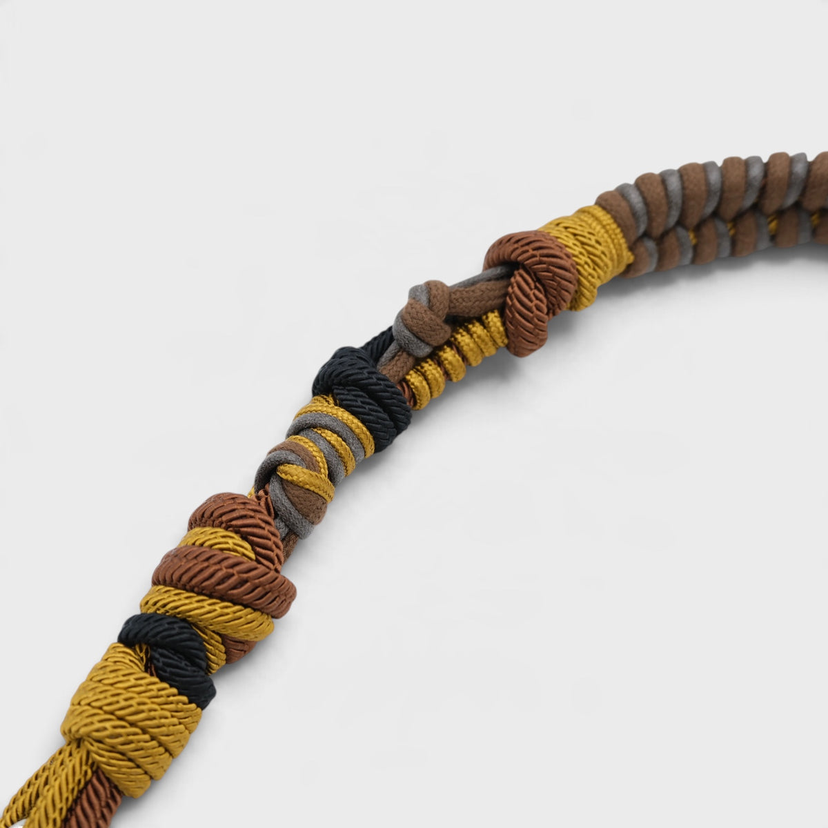 A close-up of a braided macrame shoulder strap, featuring alternating brown and gray braided sections with a small yellow accent thread, finished with a knotted top on a light gray background.