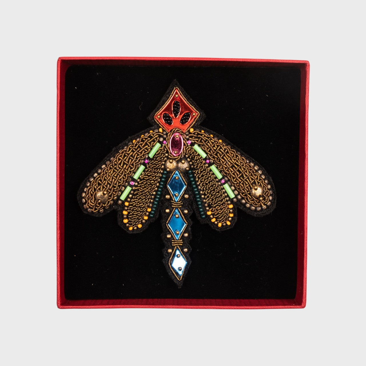 An embroidered brooch representing a dragon fly with bronze details in a box.