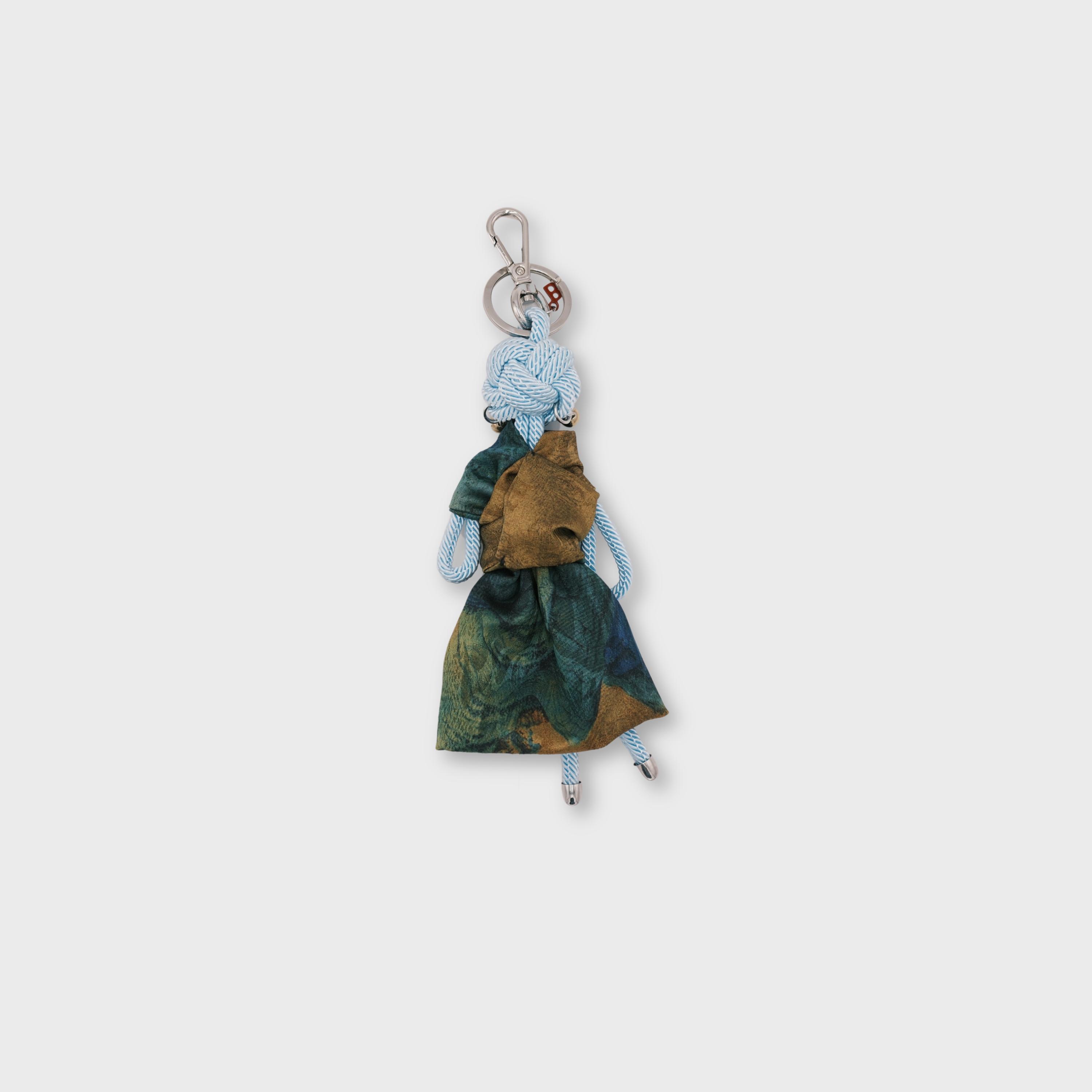 A handmade macramé keychain featuring a small light blue rope doll with a green and brown patterned dress, gold coloured earrings, and a silver metal key ring at the top.