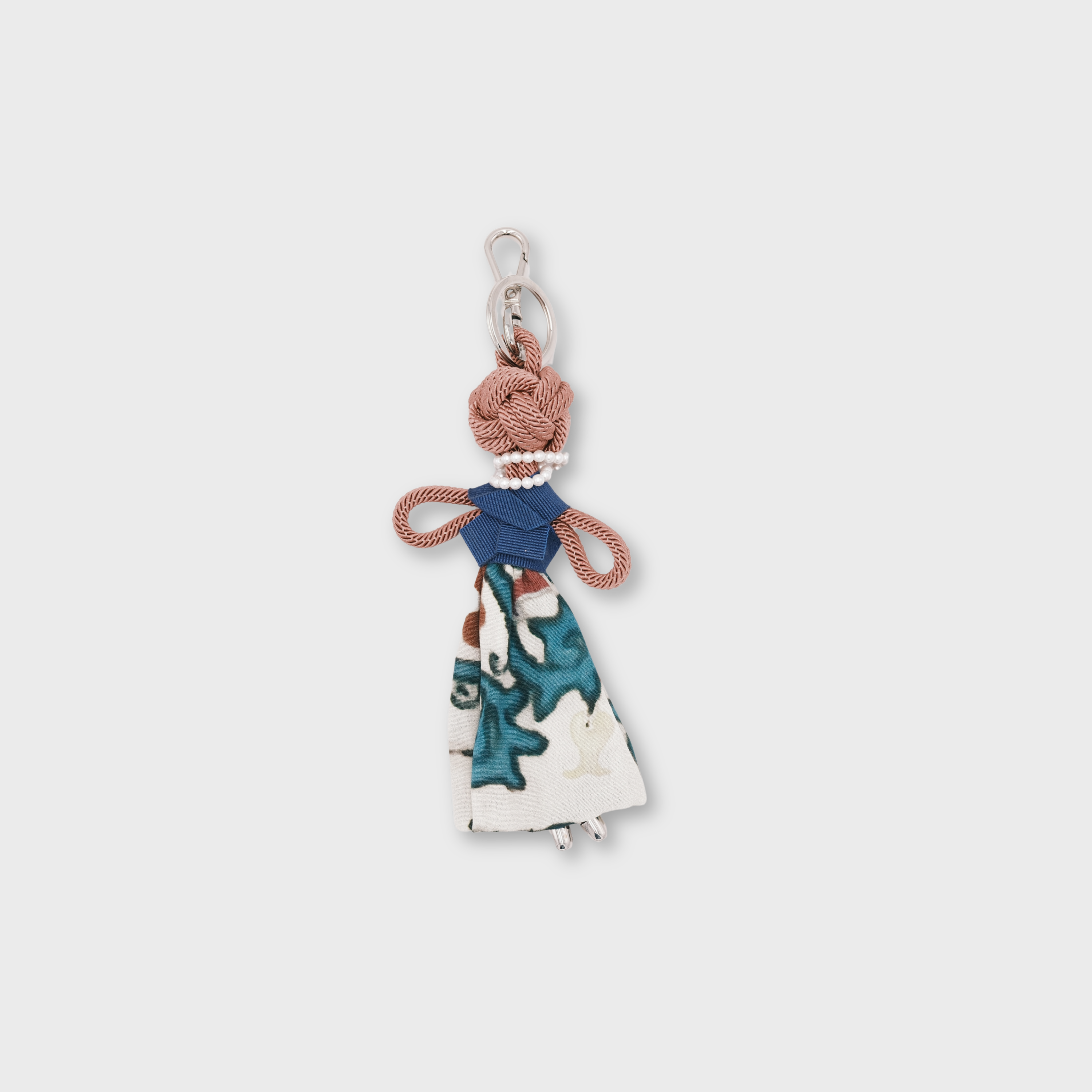 A small decorative pink macramé keychain shaped like a stylized woman, featuring a blue top, a white skirt with teal and blue abstract print, and a silver metal key ring at the top.