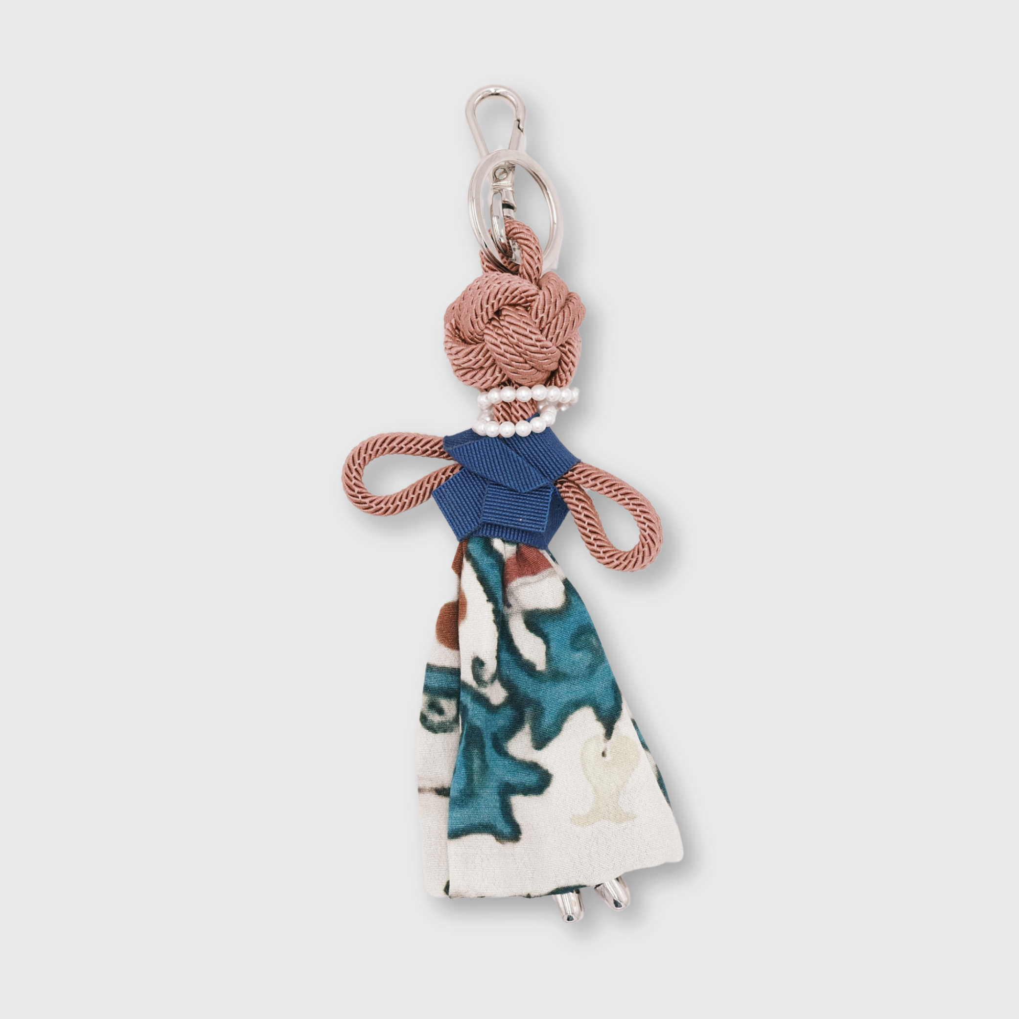 A small decorative pink macramé keychain shaped like a stylized woman, featuring a blue top, a white skirt with teal and blue abstract print, and a silver metal key ring at the top.