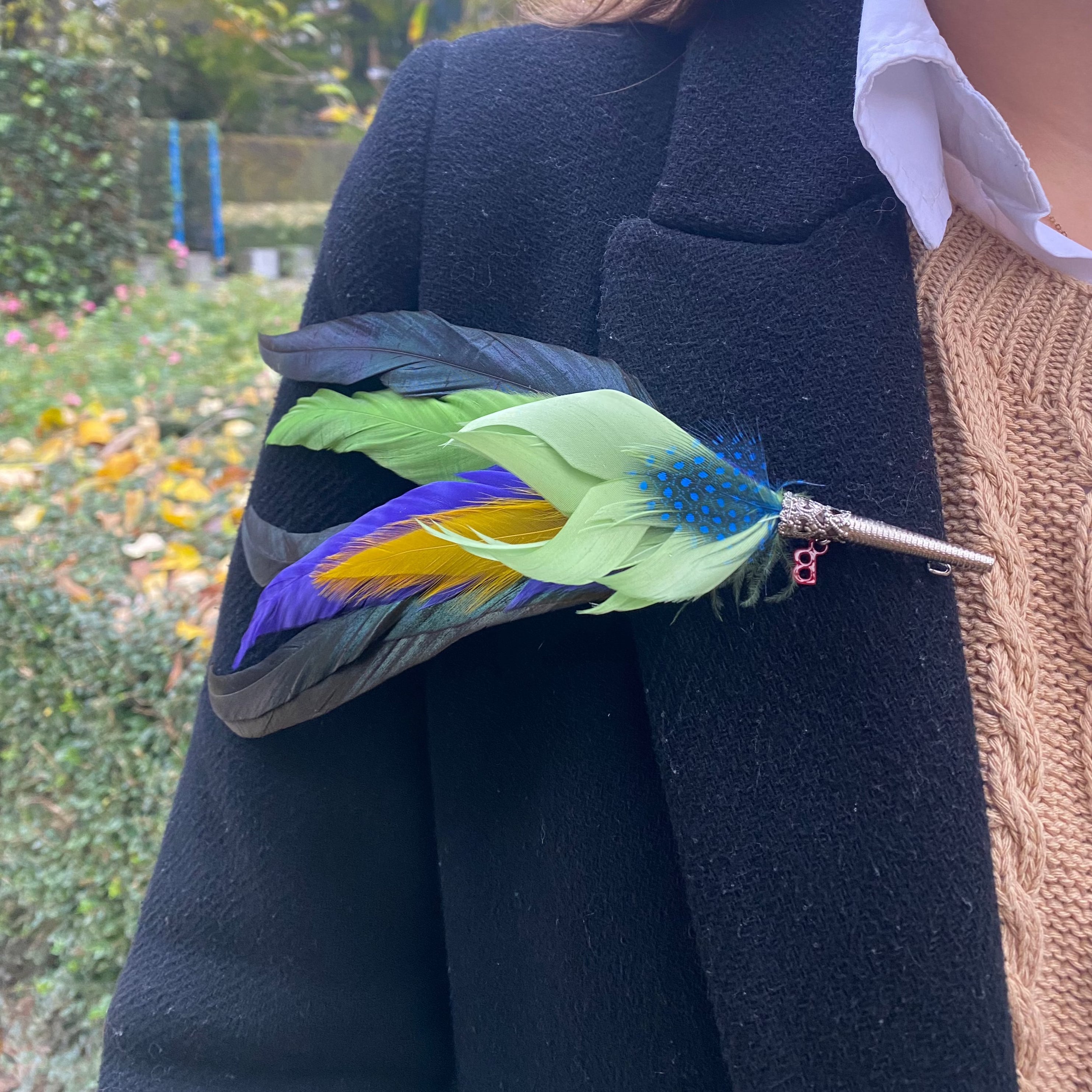 A brooch featuring dyed feathers in green, blue, and purple, attached to a metal pin and displayed on a jacket.