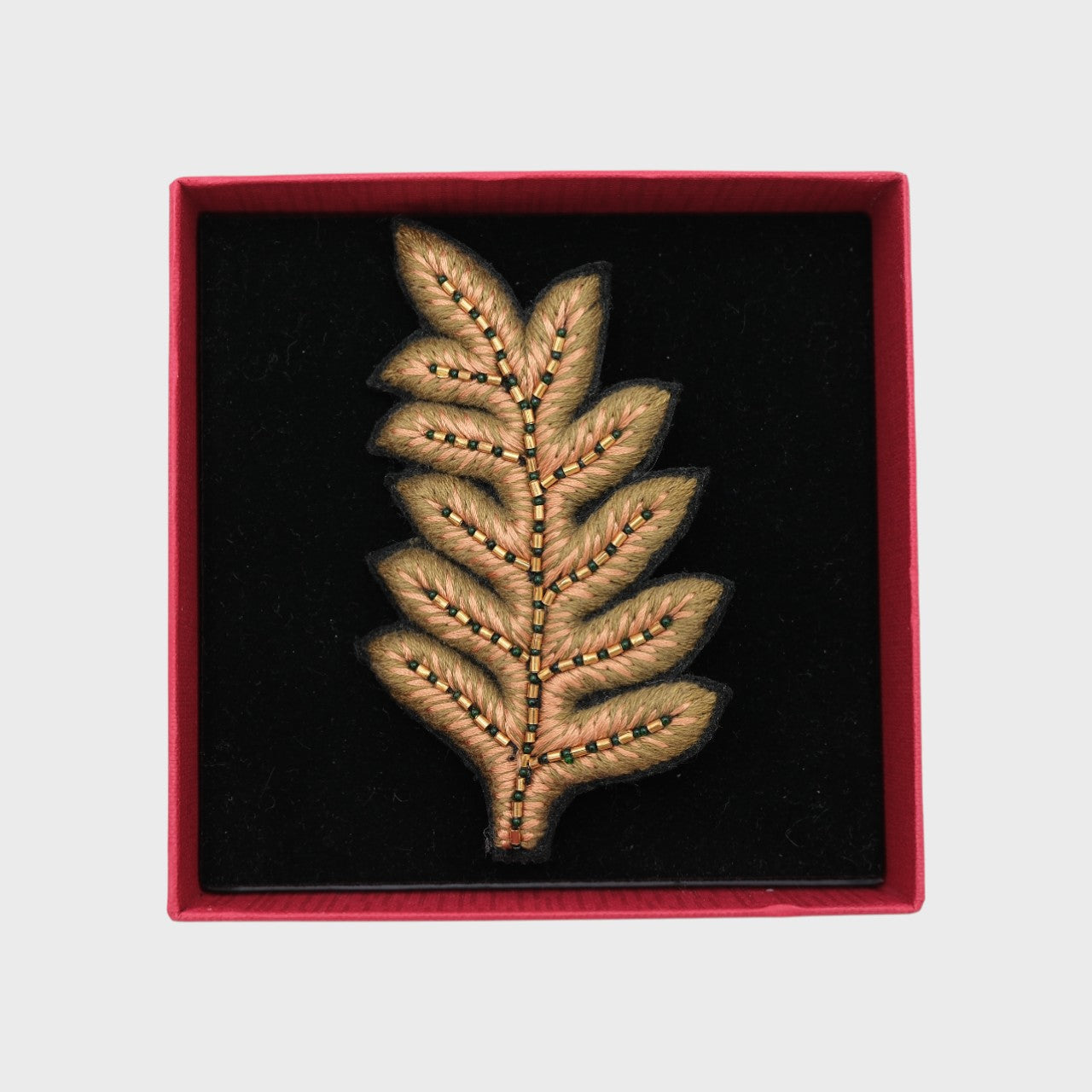 An embroidered brooch representing a fern leave in green and beige in a box.