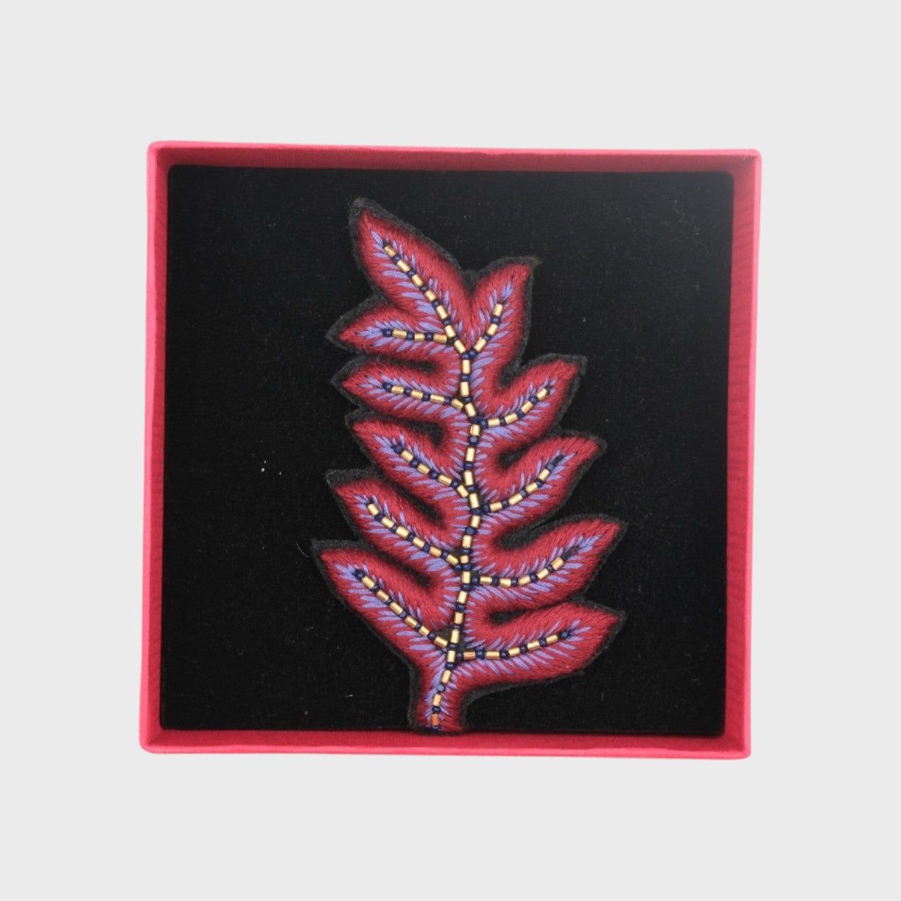 An embroidered brooch representing a fern leave in purple in a box.