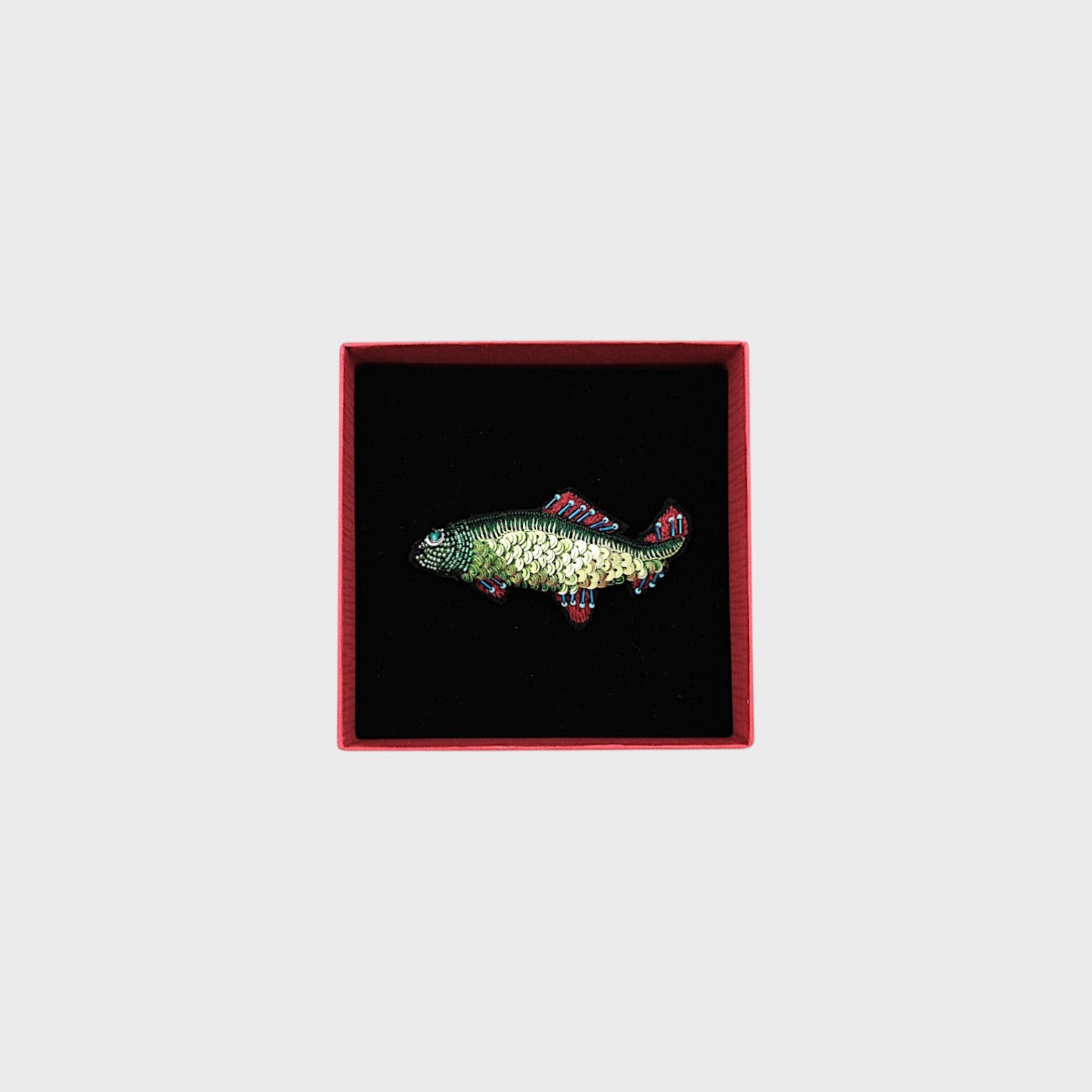 A embroidered green fish brooch woth red and blue details in a box.