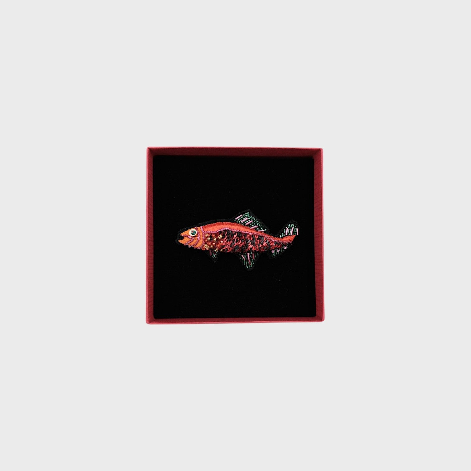 An embroidered red fish brooch with green and pink details in a box.