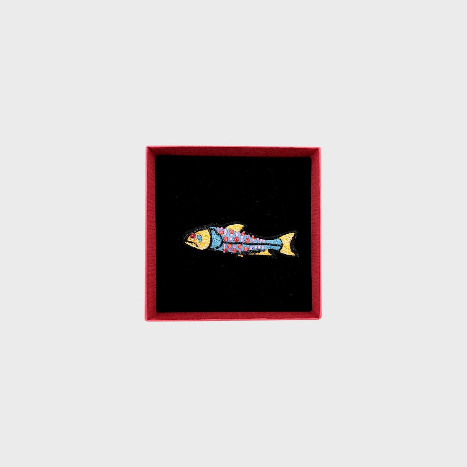 An embroidered blue fish brooch with yellow, pink and red details in a box.