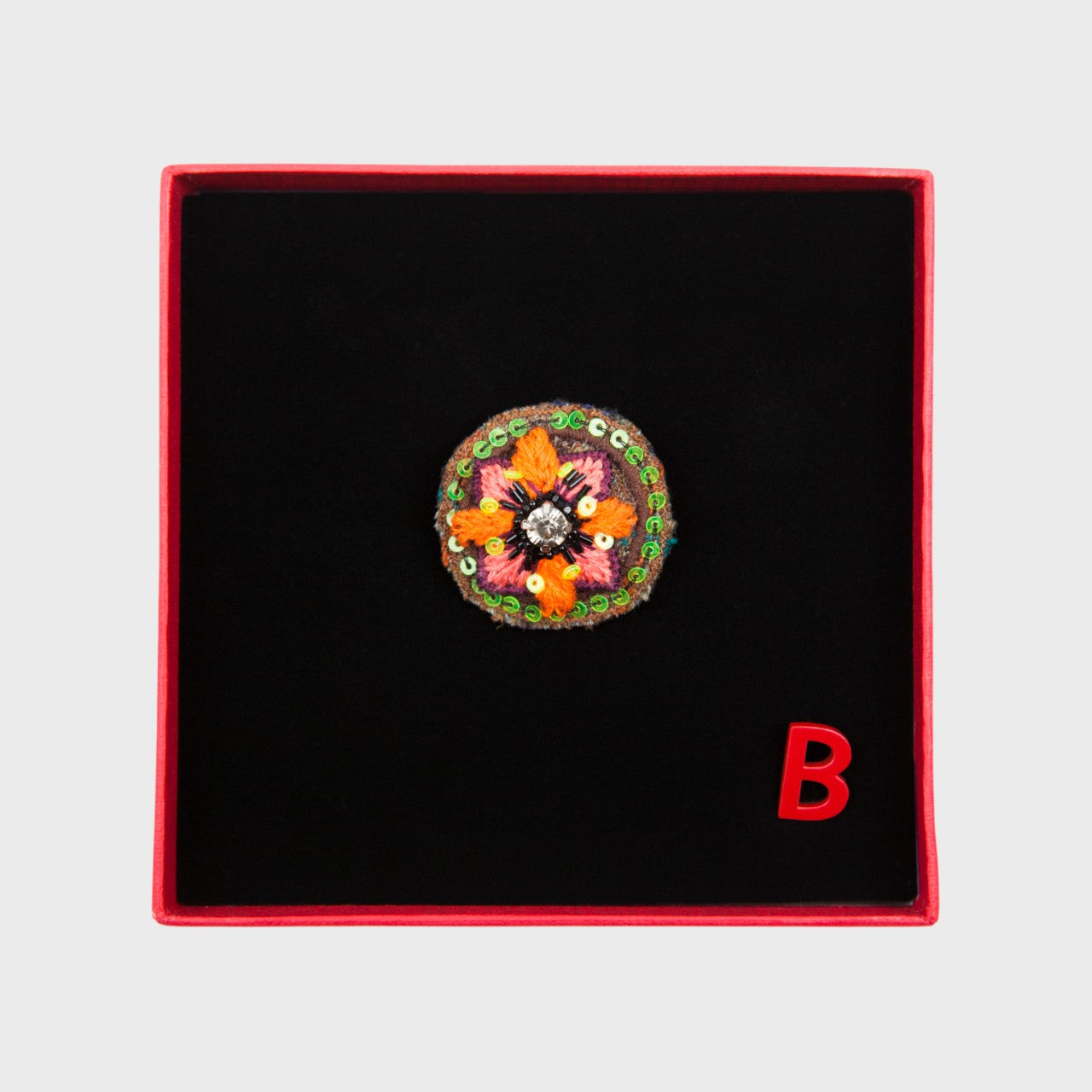 An embroidered brooch representing an orange and pink flower in a box.