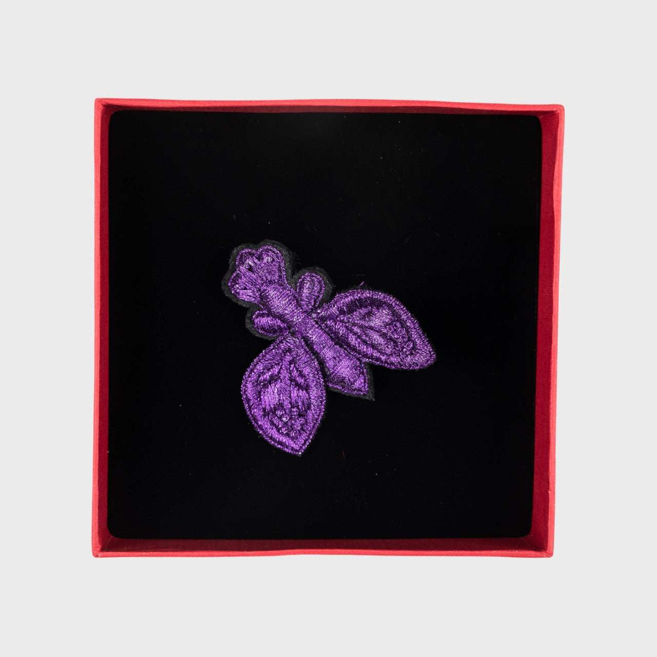 An embroidered brooch of a purple fly in a box.