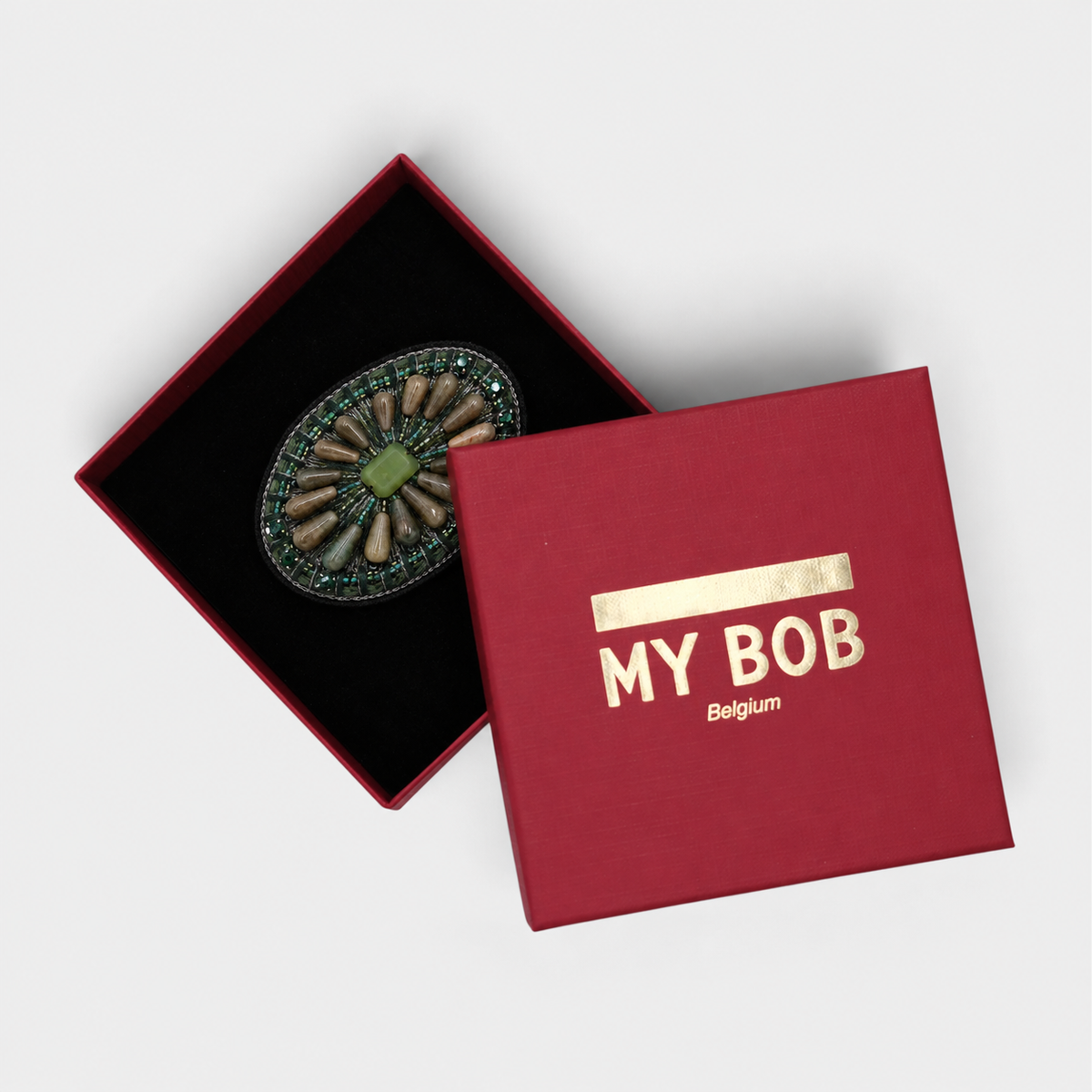 An oval brooch with a green central stone and natural petal-like accents, displayed inside a red presentation box with a black velvet lining on a light gray background.