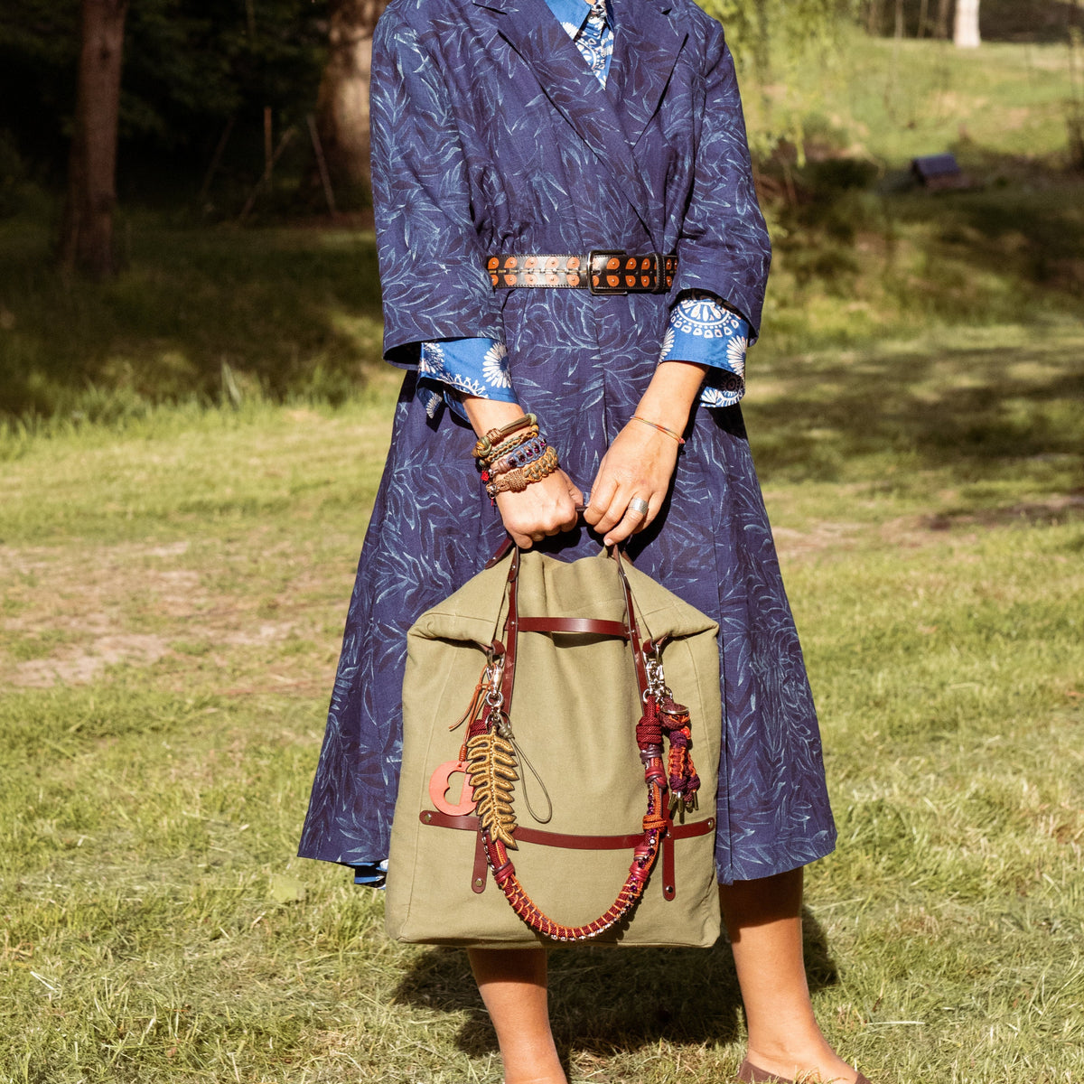 Woman in a blue outfit with a hat holding a bag with a green embroidered fern leave bag char on it, on a grassy field