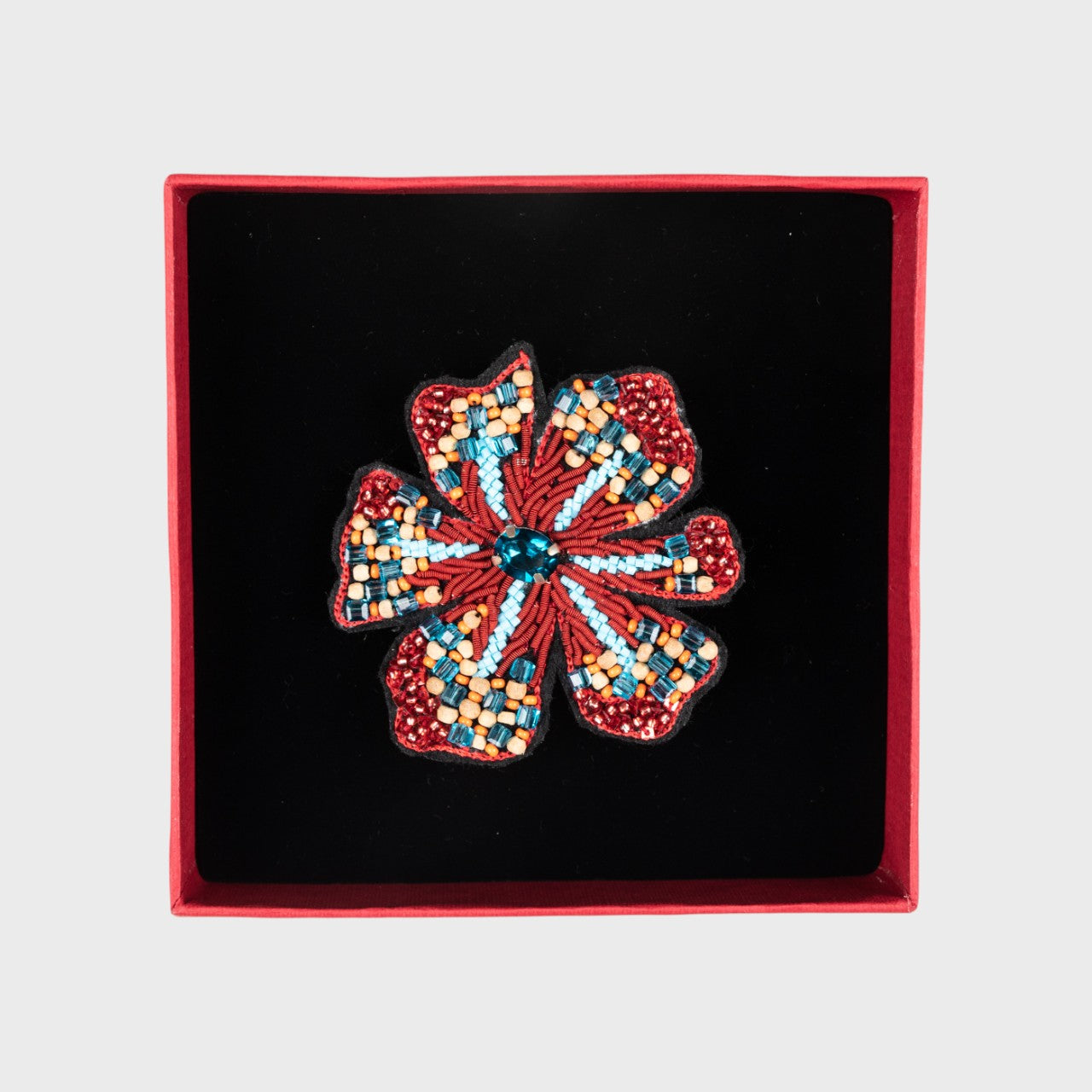 An embroidered brooch representing a flower with red details in a box.