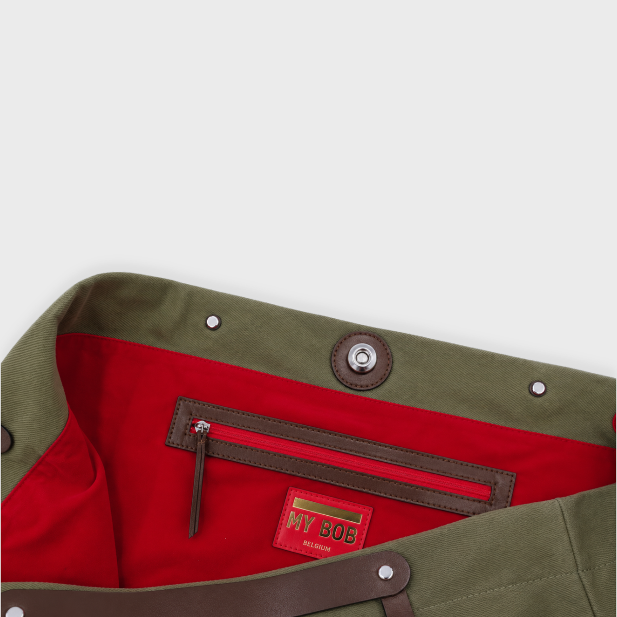 Close-up of the interior waistband of an olive green canvas bag, showing a red inner lining, brown leather trim, metal rivets, and a red brand patch reading 'MY BOB'.