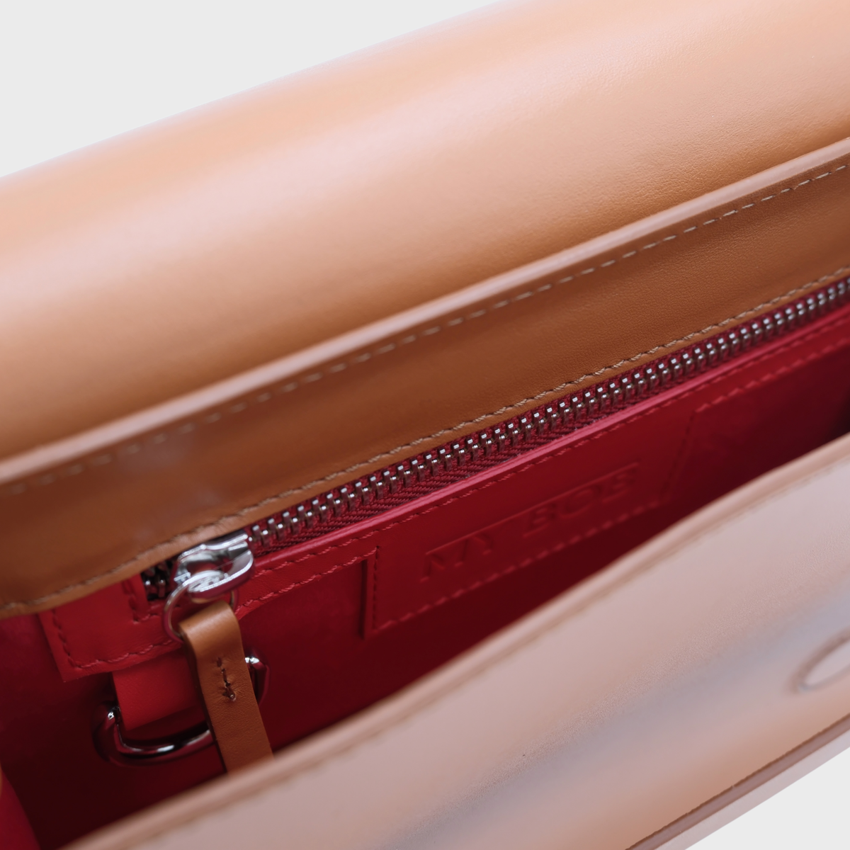 Close-up of a beige leather handbag interior with a red zippered pocket and silver hardware, showing a small embossed brand logo MY BOB on the red lining.