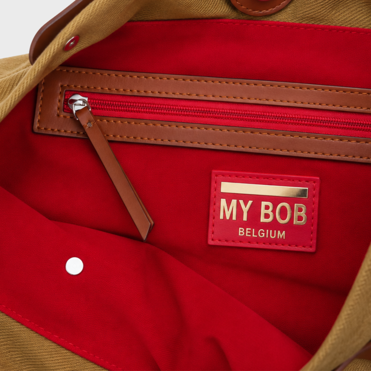 Close-up of the interior of a bag showing a bright red fabric lining, tan leather trim, a zippered pocket, and a red patch with gold lettering that reads 'MY BOB BELGIUM'.