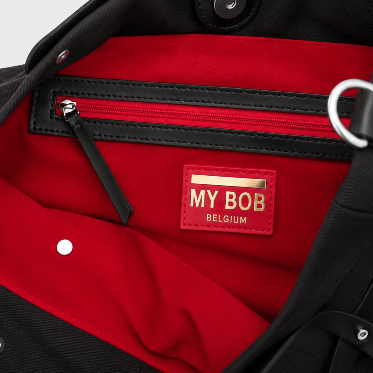 Close-up of the interior of a black bag with a bright red lining, featuring a red zipper pocket, a red brand patch reading 'MY BOB BELGIUM' with gold lettering, and metal hardware.