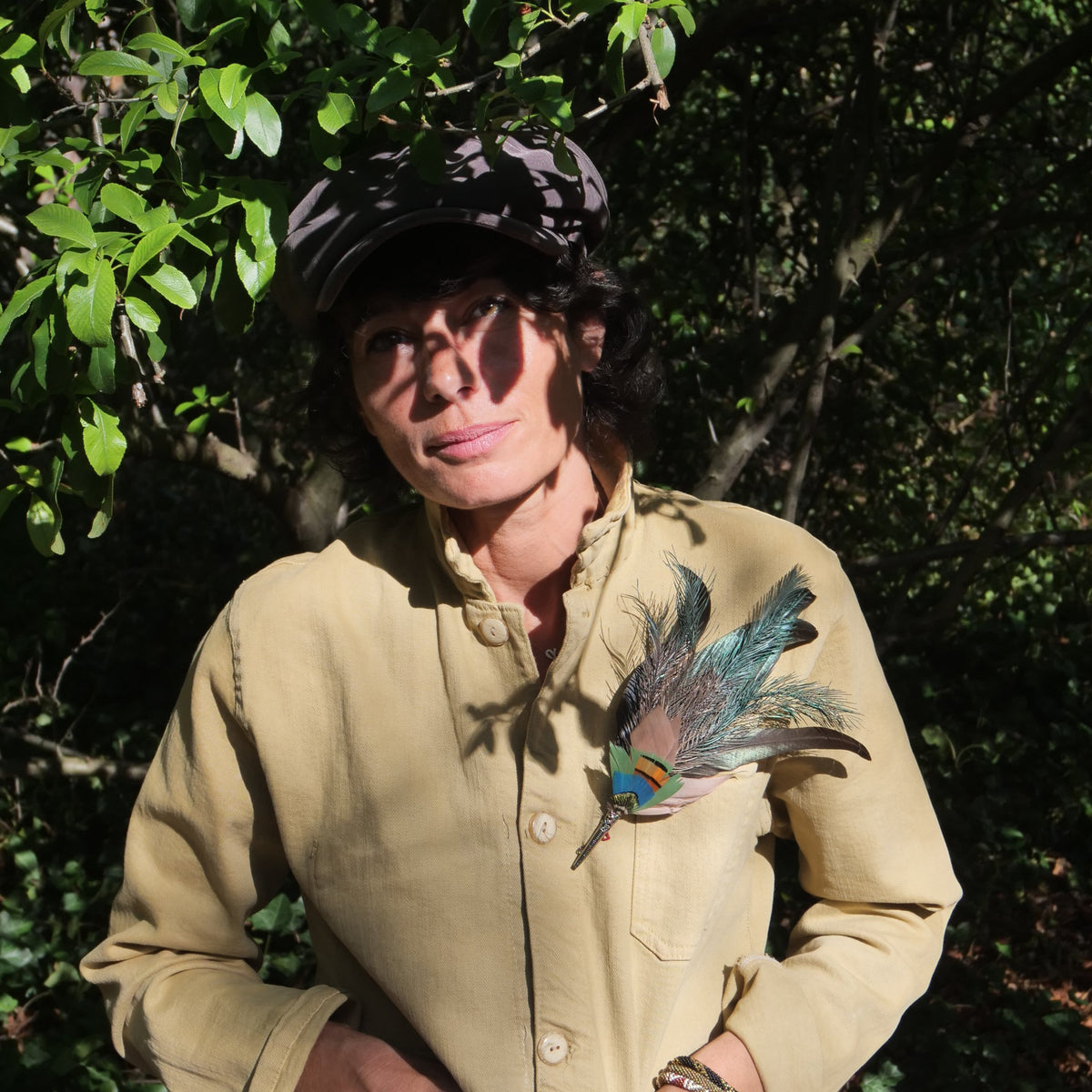 A person wearing a beige jacket and a black hat adorned with a multicolored feather brooch, standing outdoors with foliage in the background.
