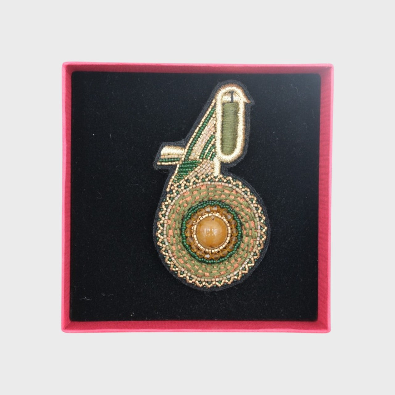 An embroidered brooch representing a green kiwi bird in a box.