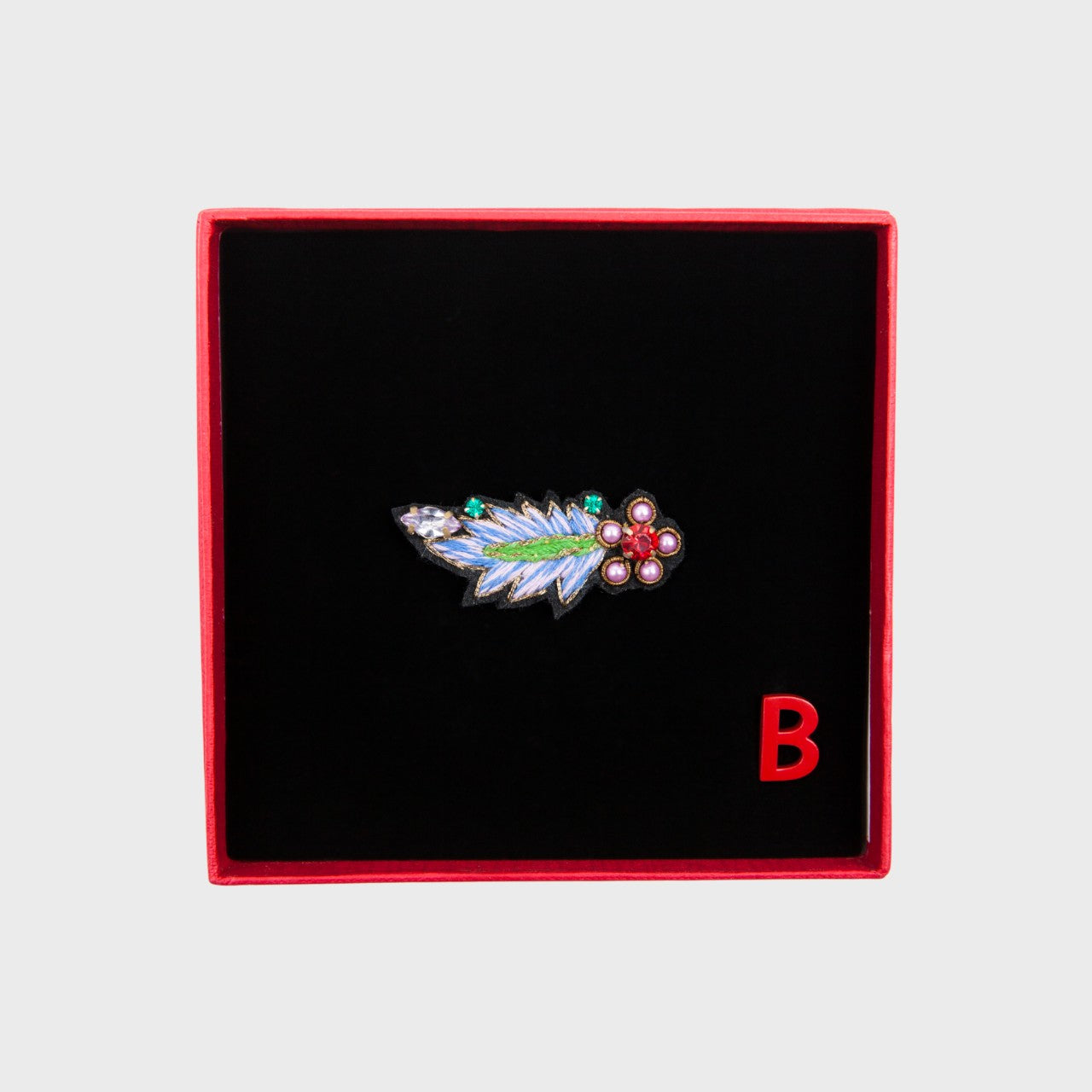 An embroidered brooch representing a blue leaf in a box.