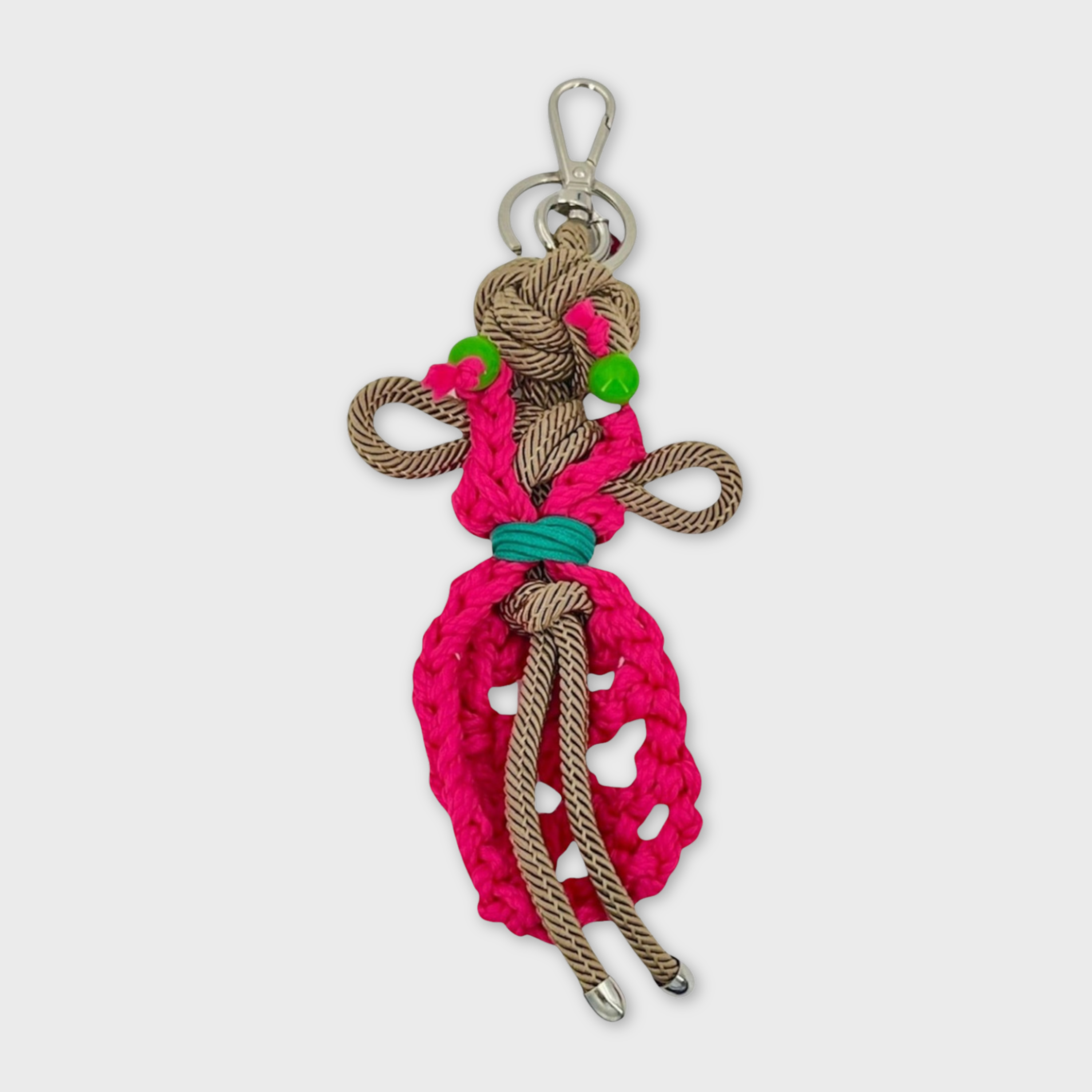 Handmade tan macramé bag charm featuring a female character, meant to represent a colourful dancer wearing a long pink dress and green earrings. She is attached to a silver clasp.