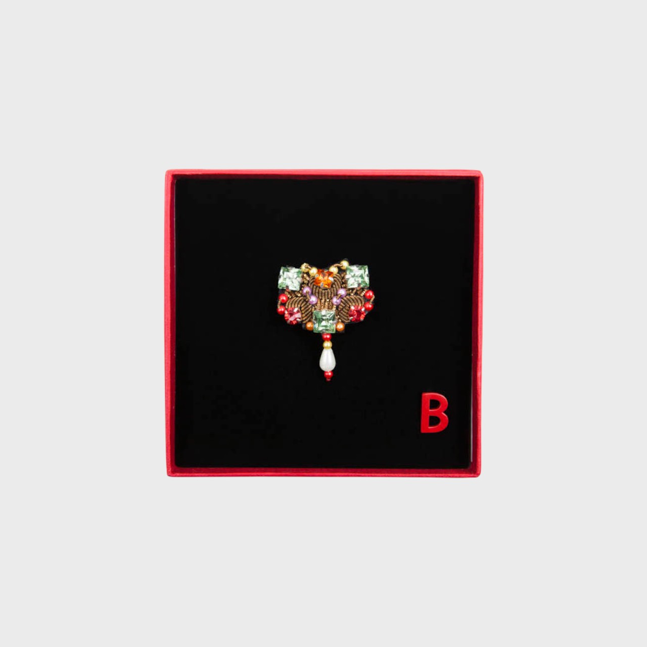 An embroidered brooch with green stones and bronze details in a box.