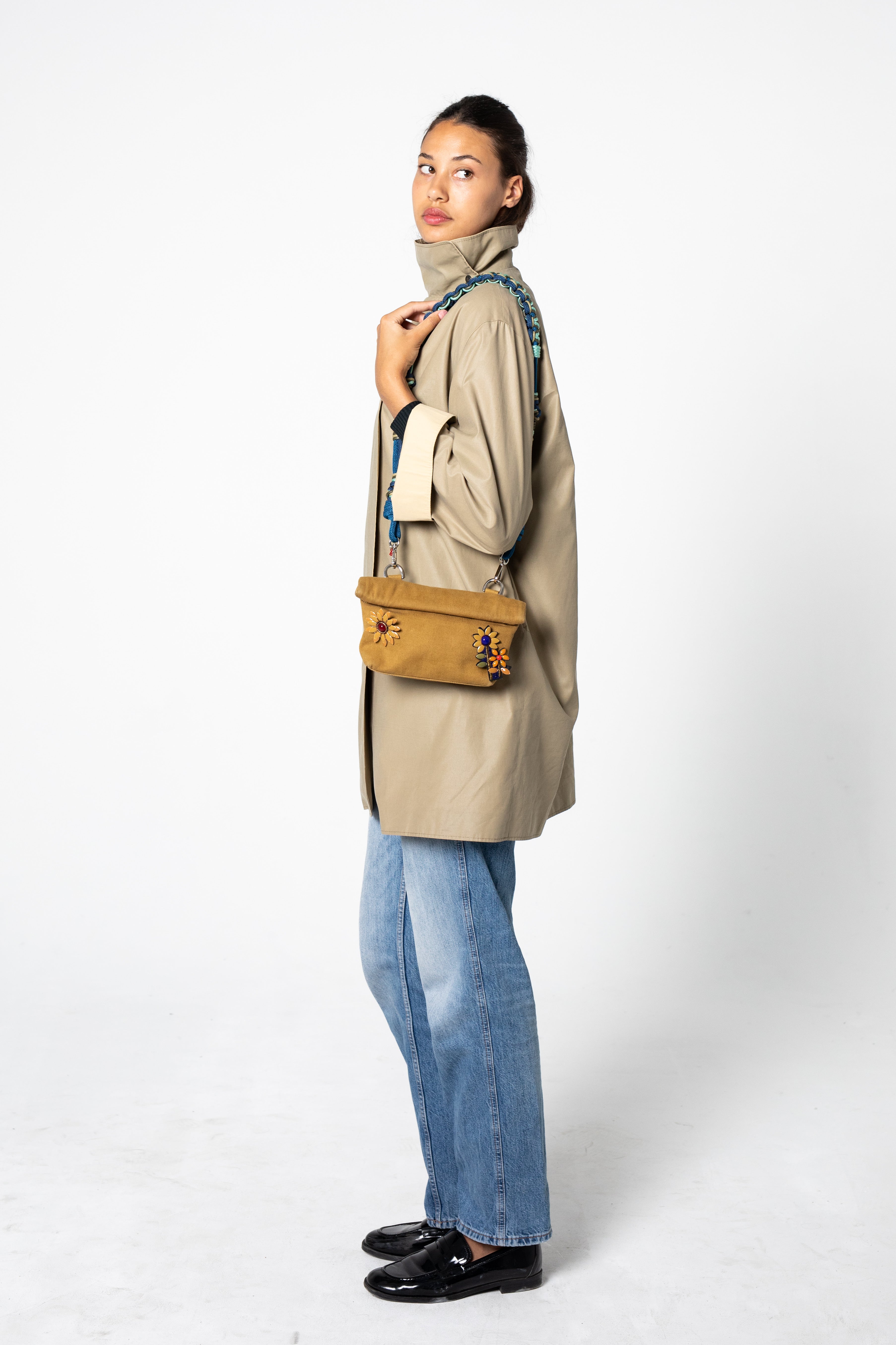 Woman wearing a beige coat and blue jeans, holding a yellow MINOPTI bag and a blue ALICE bag strap against a white background