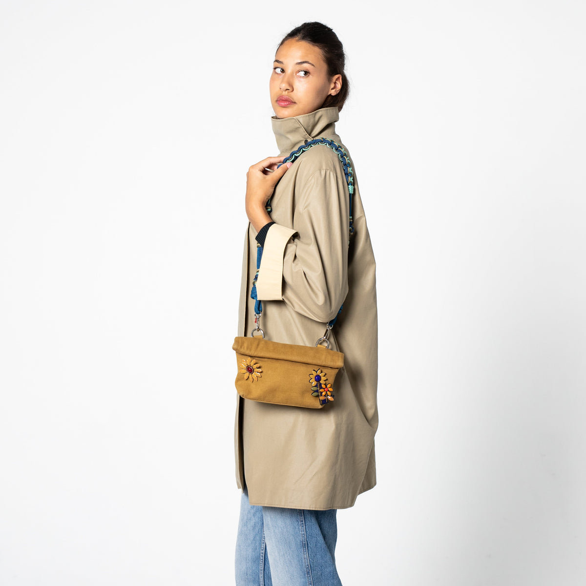 Woman wearing a beige coat and blue jeans, holding a yellow MINOPTI bag and a blue ALICE bag strap against a white background