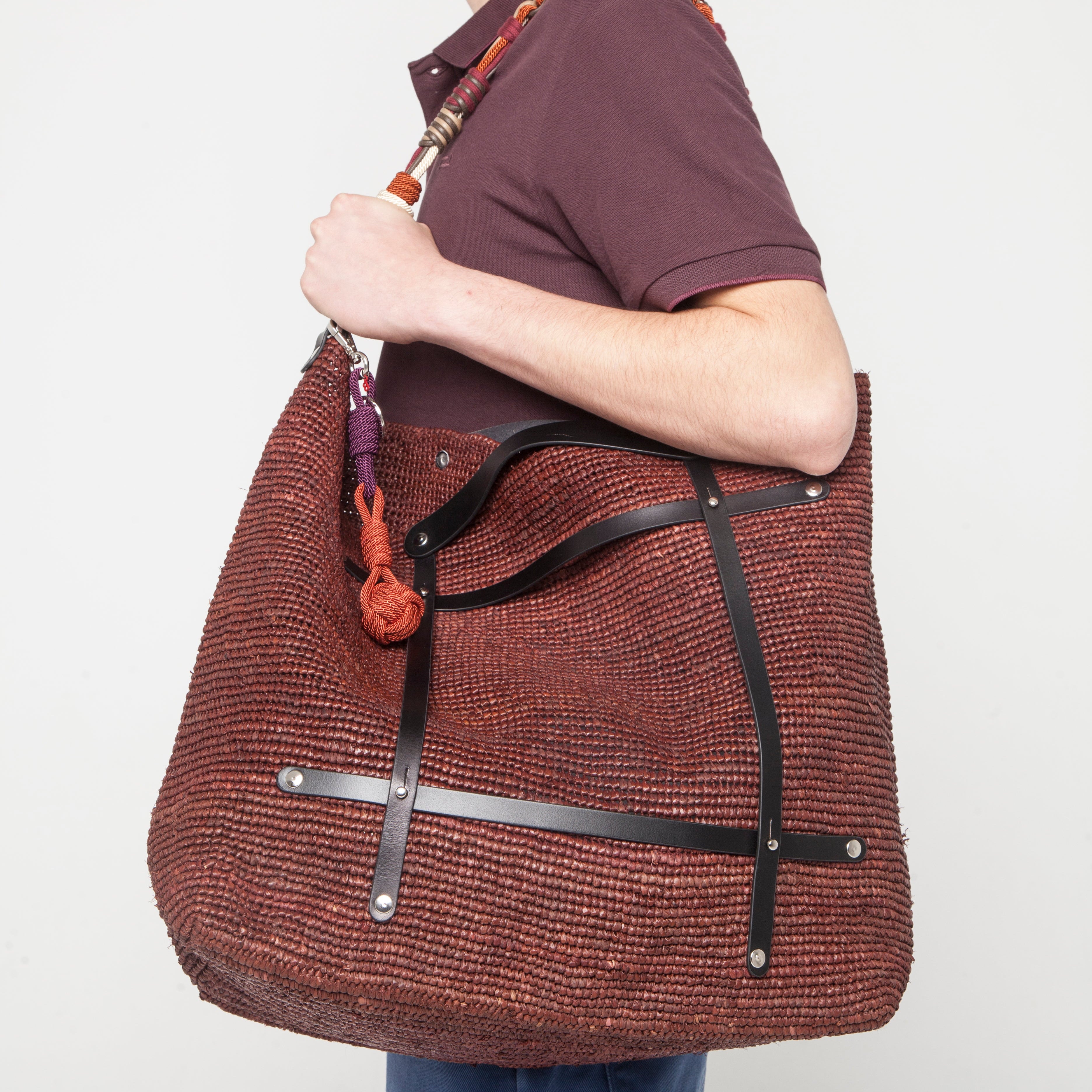 A man in a dark brown polo shirt and blue jeans is shown from the side, carrying a brown raffia candide weekender bag with black leather harness, a decorative orange sailorball bag charm and an colorful Alice shoulder strap in orange and red.