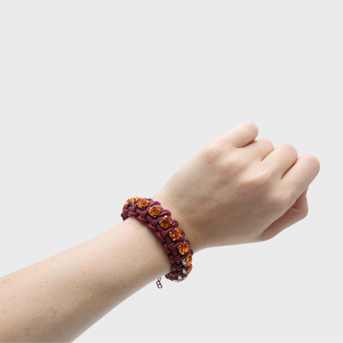 A close-up of a person's wrist wearing a purple woven macrame bracelet adorned with amber-colored oval stones, set against a plain light gray background.