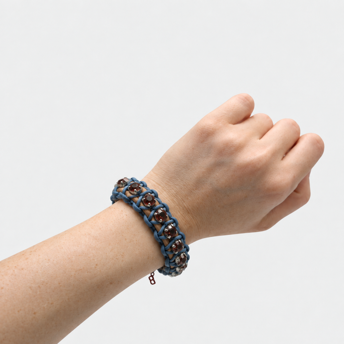A close-up of a person's wrist wearing a blue and brown woven bracelet with small dark purple beads, shown against a plain light gray background.