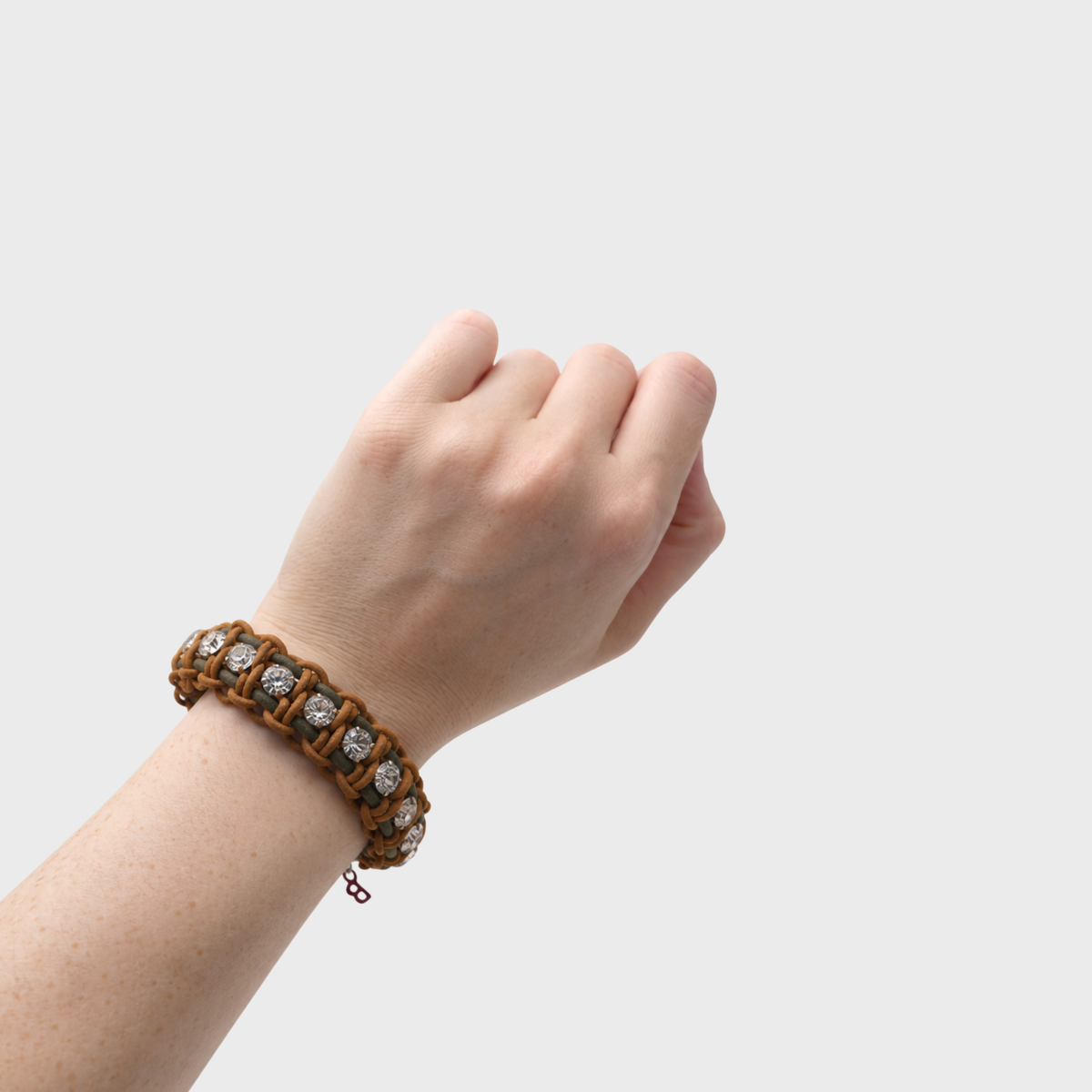 A close-up of a person's wrist wearing a brown woven bracelet with clear gemstones set in a chain link design, against a light gray background.